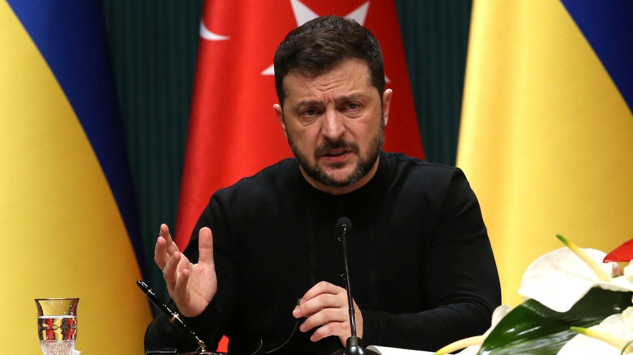 Volodymyr Zelensky gestures with his hands as he speaks at a press conference. There's a microphone and flowers on the table in front of him, and behind him the Ukrainian and Turkish flags. He's wearing a black long sleeeved t-shirt.