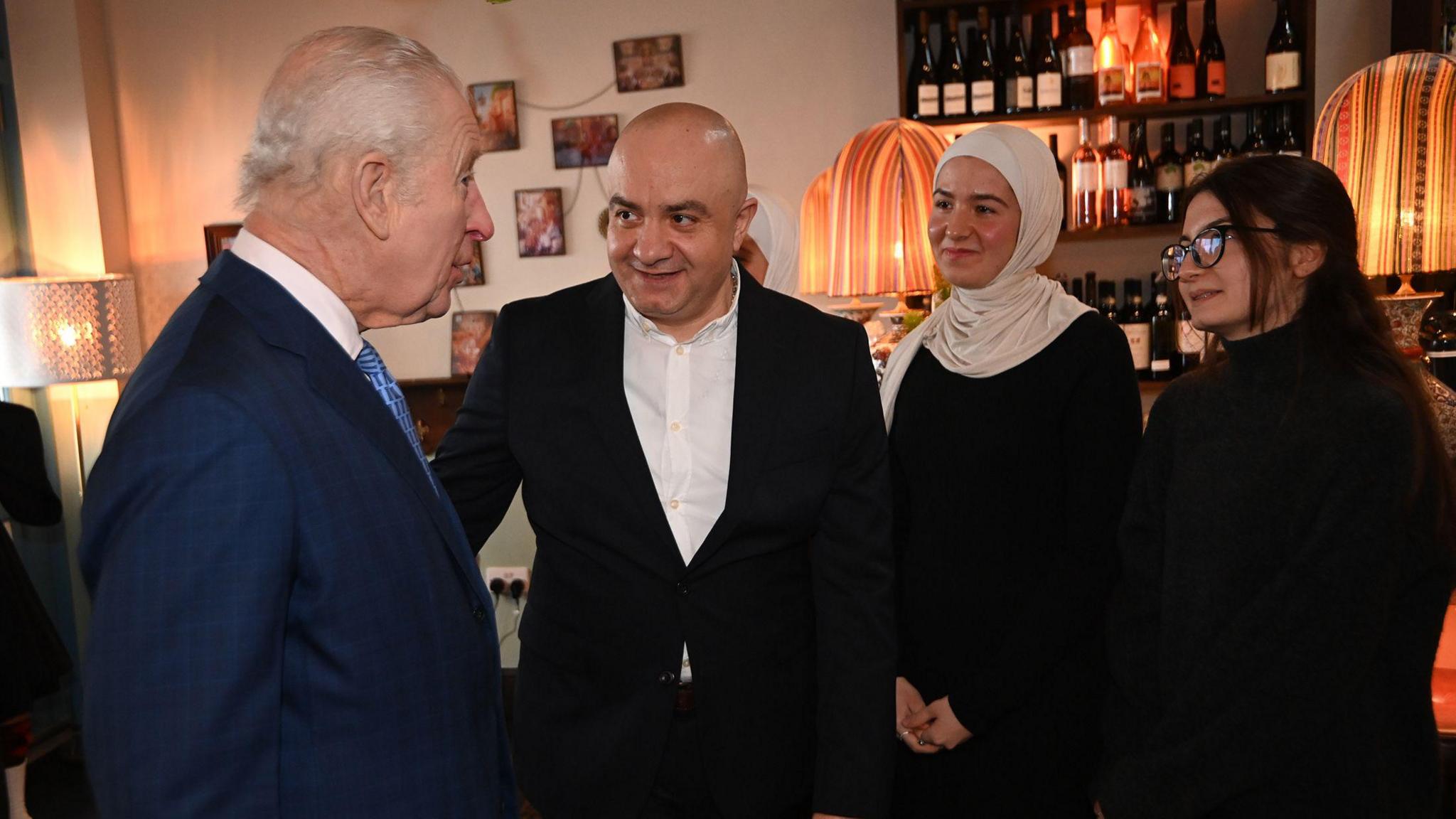 King Charles standing by another man and two women, in conversation. He is wearing a navy suit and blue tie. 