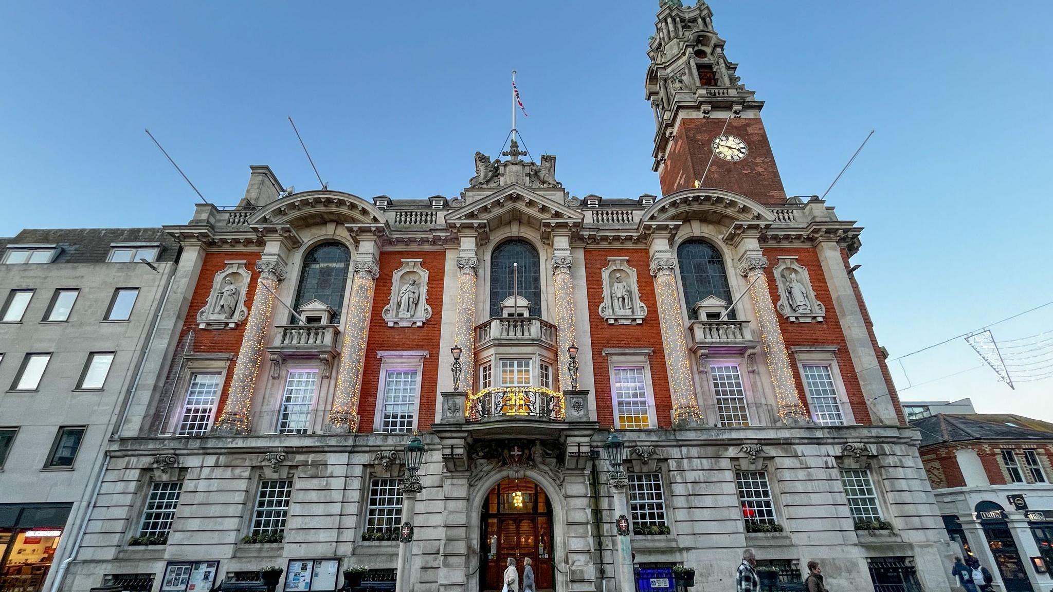 A large ornate building made of grey stone with red brick sections and statues embedded into the walls. There is a large door at the front and a balcony above it. There are a number of flag poles suspended from balconies