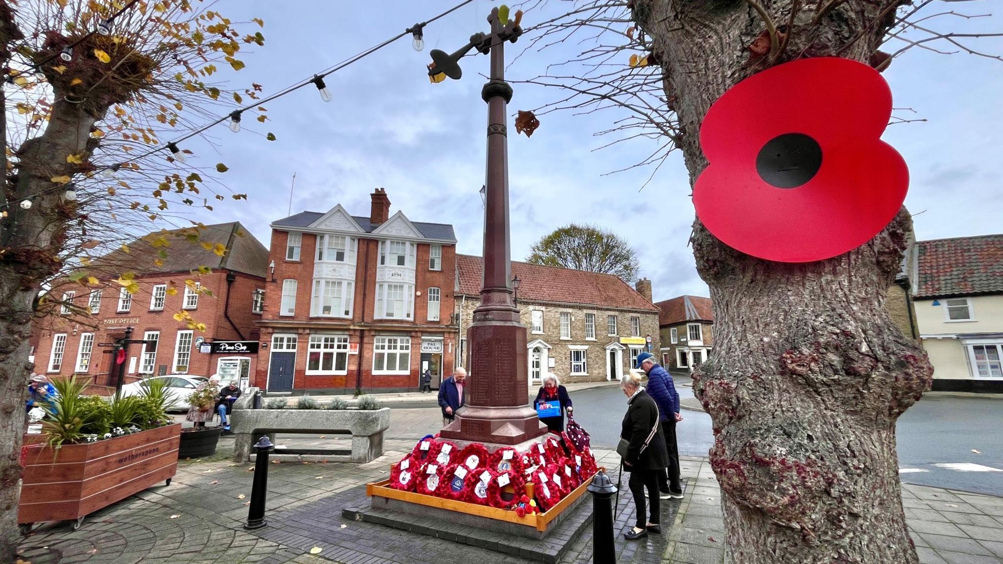 A large red poppy is pinned to a tree in the foreground. In the background a small group of people gather around a tall war memorial in a town centre. It has lots of poppy wreaths at its base.