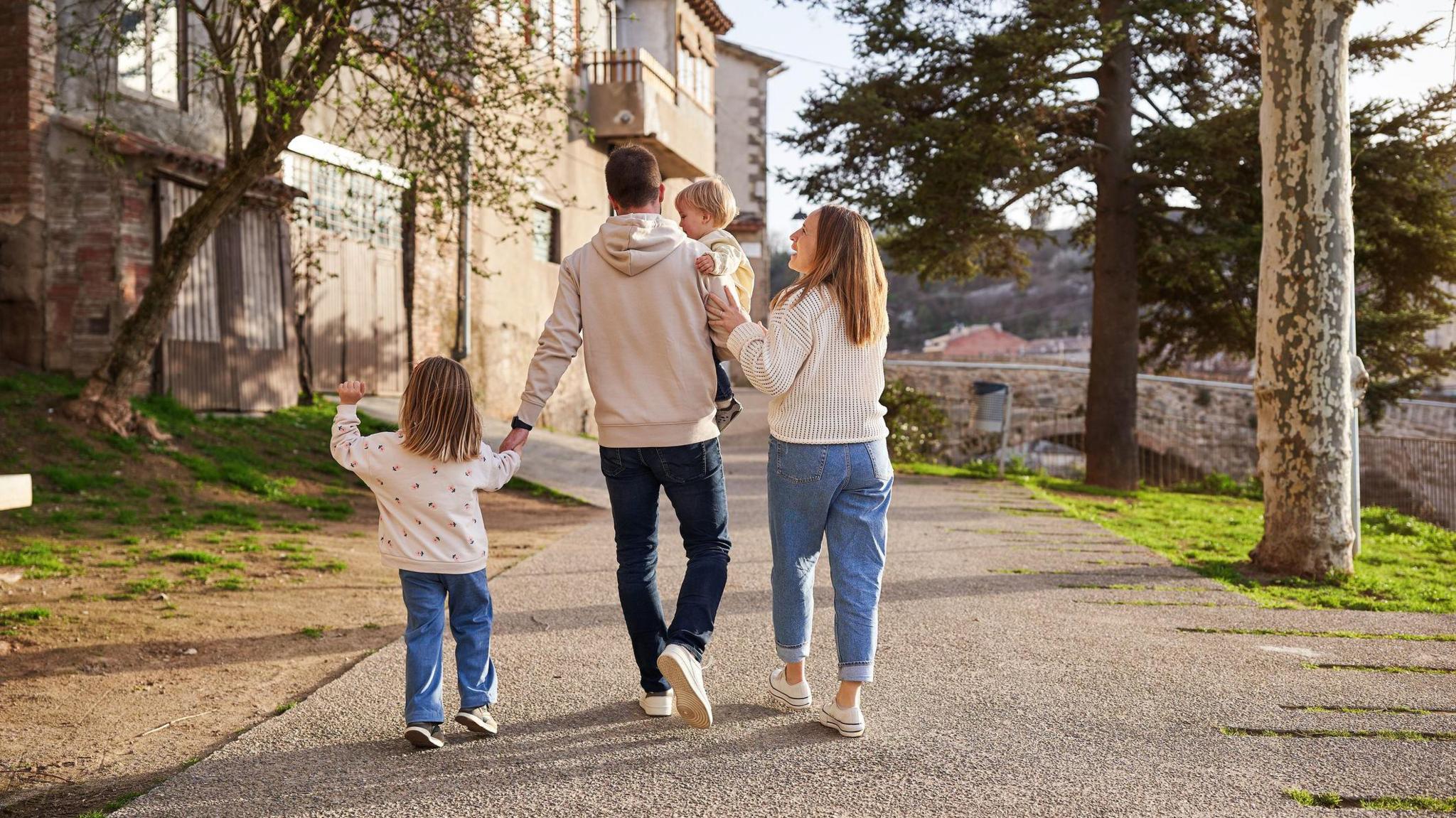 A family of four - two adults and two young children - walking down a path. There is an old building on the left and a bridge in the distance on the right. The man is wearing a cream hoodie and blue jeans with white shoes. The man is holding a child. The child to his left is wearing a white jumper with pink flowers on, blue jeans and shoes. She is holding his hand. The woman is standing to the right of a man and is wearing a white crochet jumper, light blue jeans and shoes.