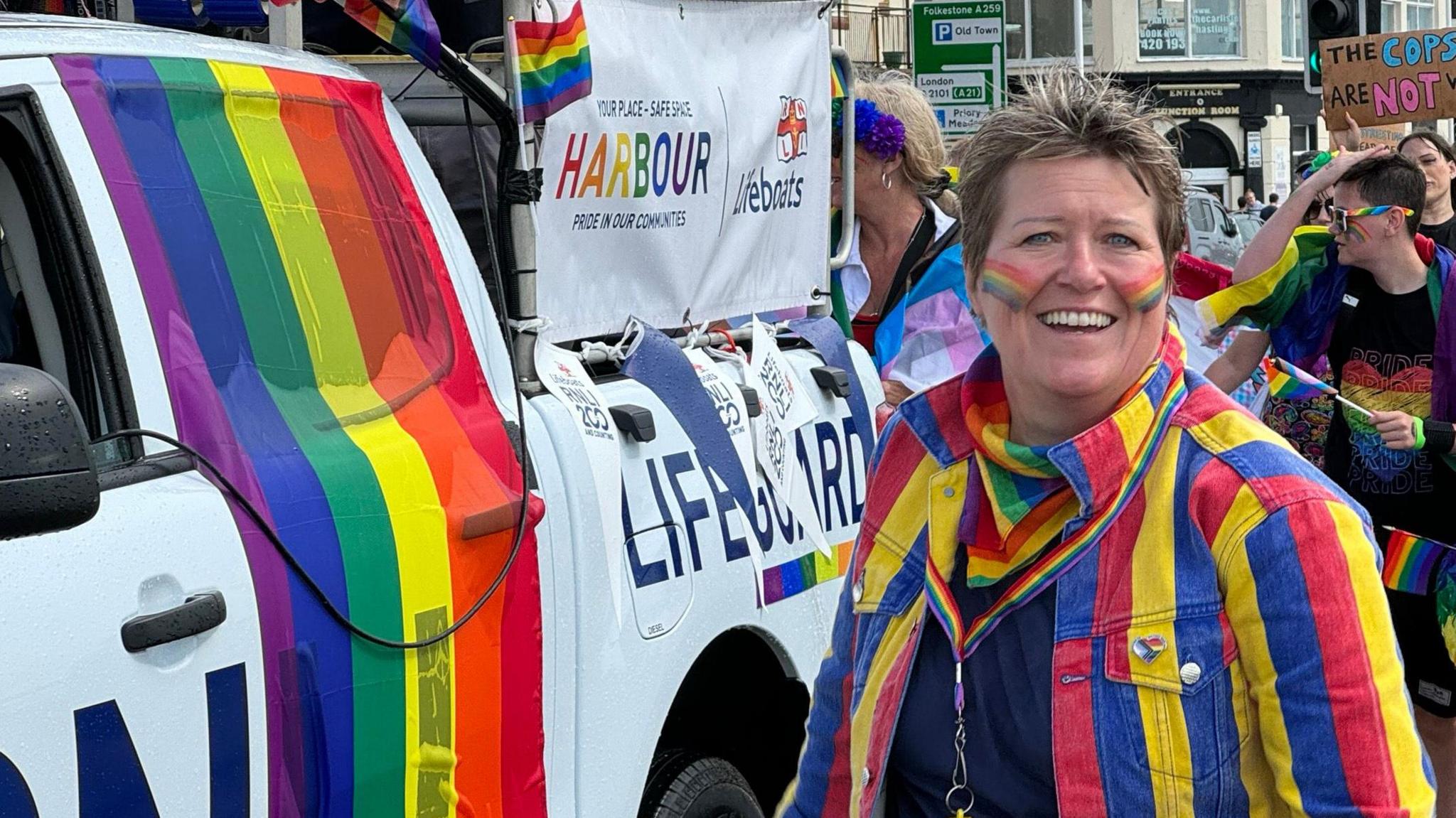 A parade with a smiling person looking at the camera in the foreground in a stripy jacket while in the background a lifeboat truck is decorated in a rainbow flag