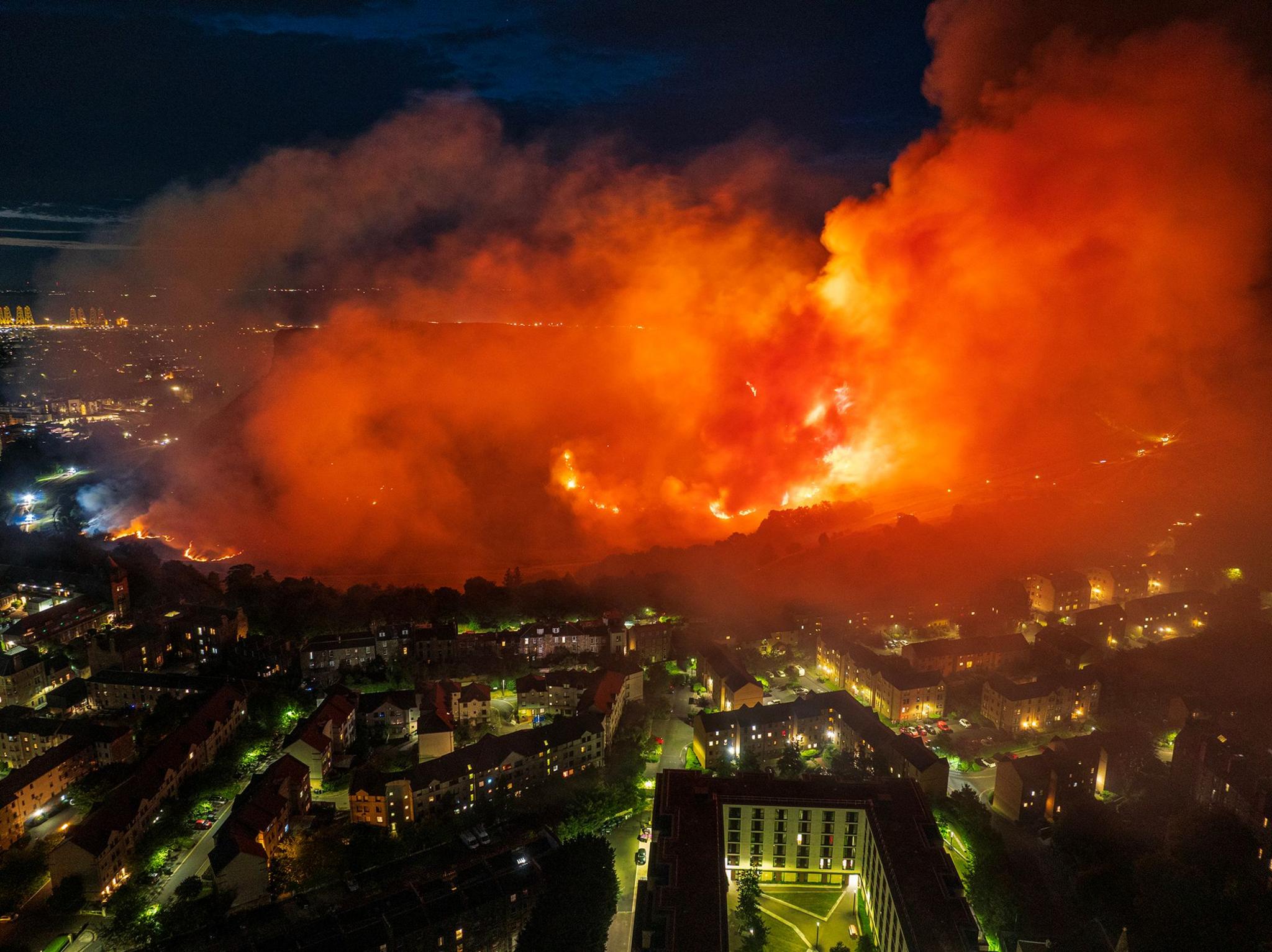An aerial shot on Arthur's Seat burning. There are orange flames and large plumes of smoke.