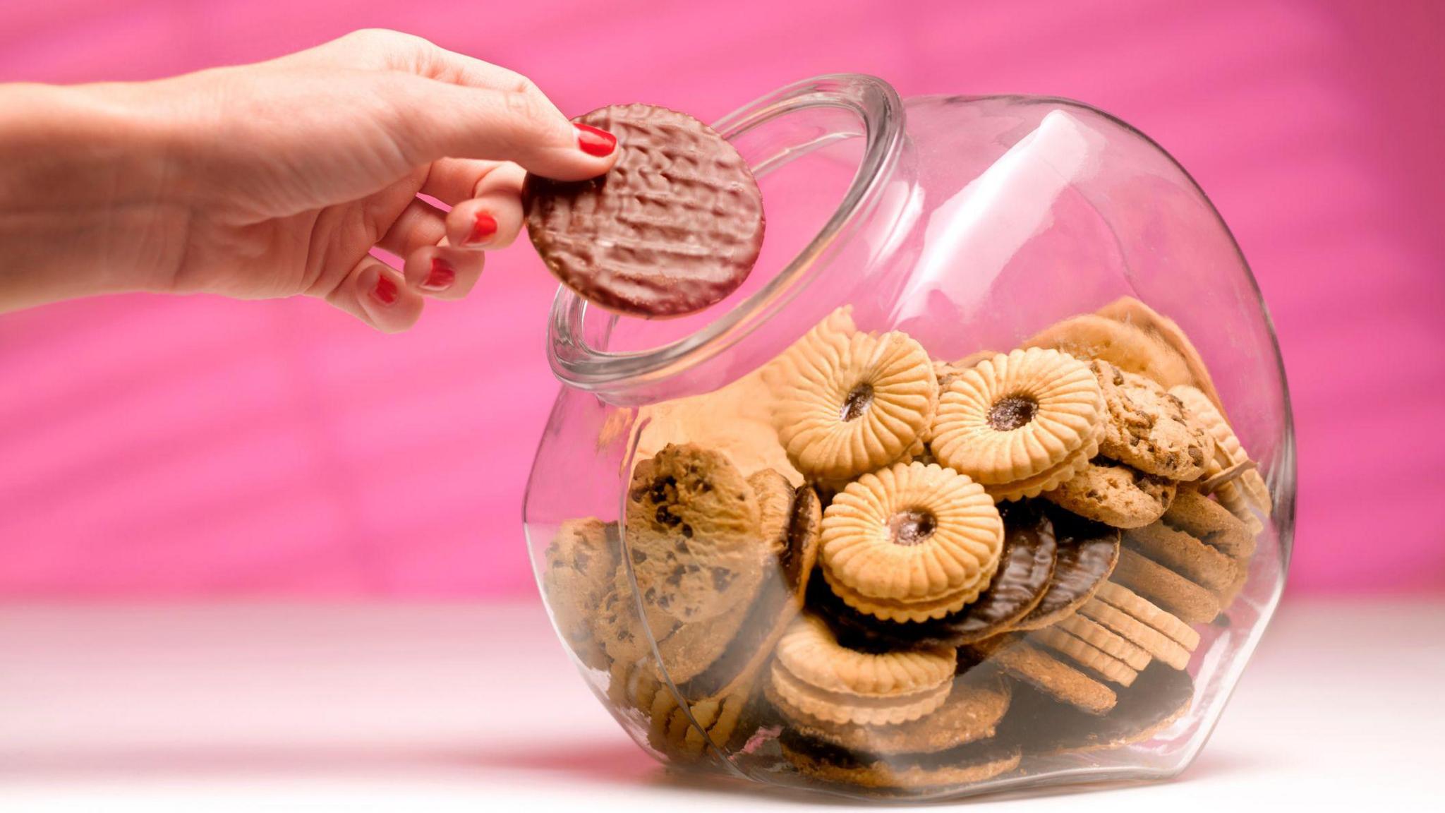 A clear biscuit tin with biscuits in it. A hand is reaching in and picking up 