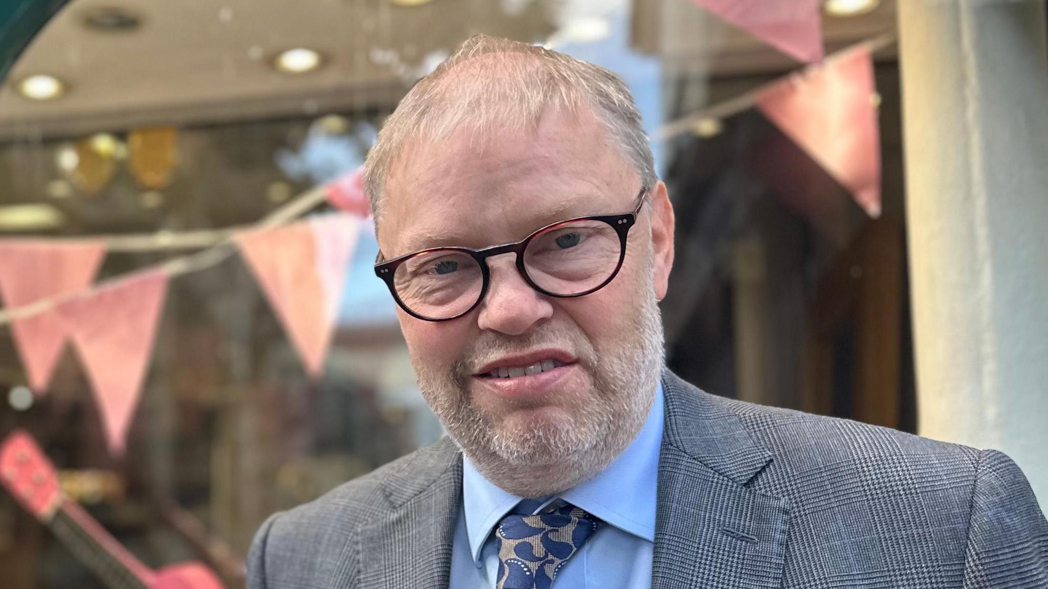 Christopher Hardman smiles at the camera. He has thinning grey hair and wears glasses as well as a grey suit, blue shirt and tie. 