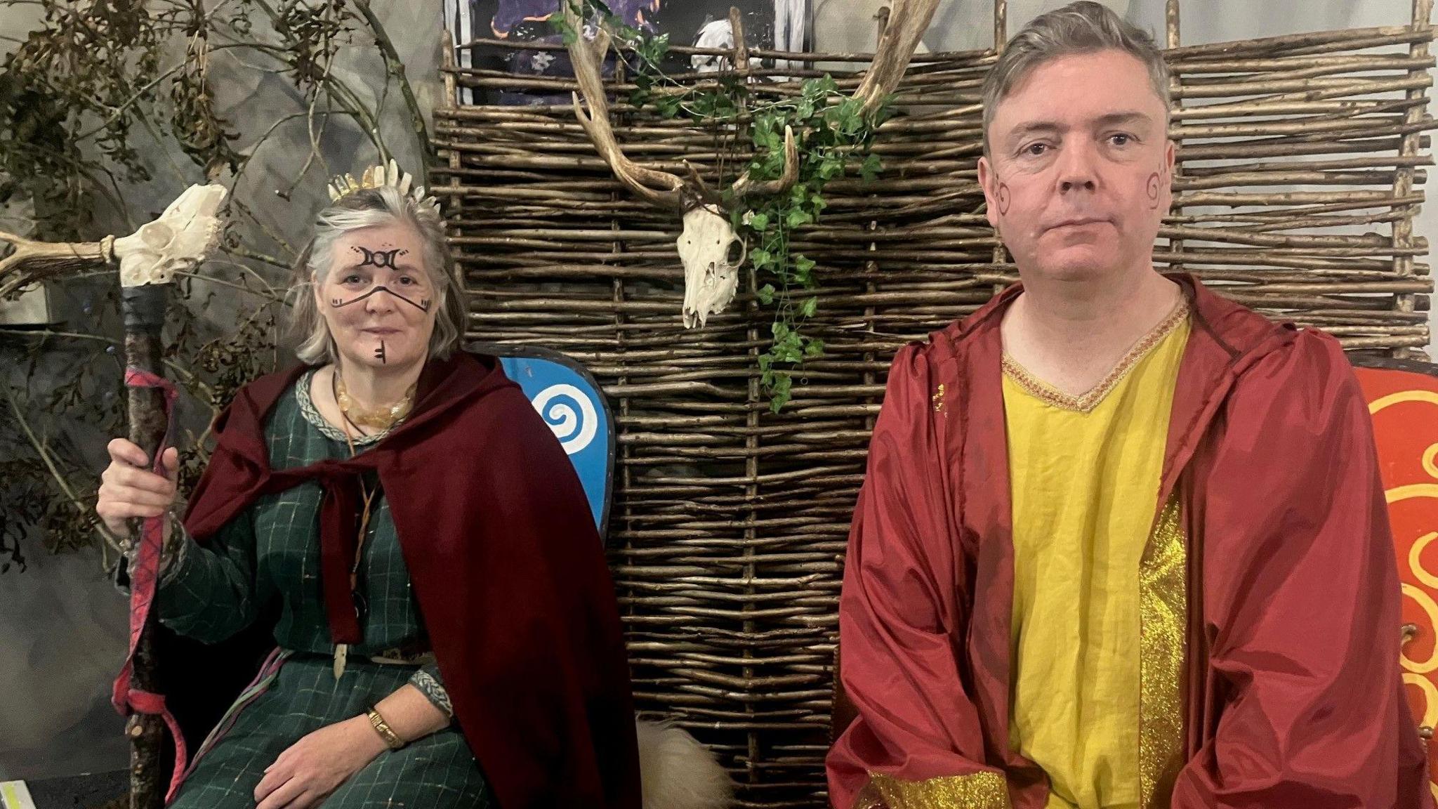 Carolyn has a staff in her hand with a bone design at the top. She has patterns in black painted on her face. She's wearing a green dress and a maroon cape. Alan is wearing a yellow costume with a maroon coat with gold edges. There's an animal's skull behind them.