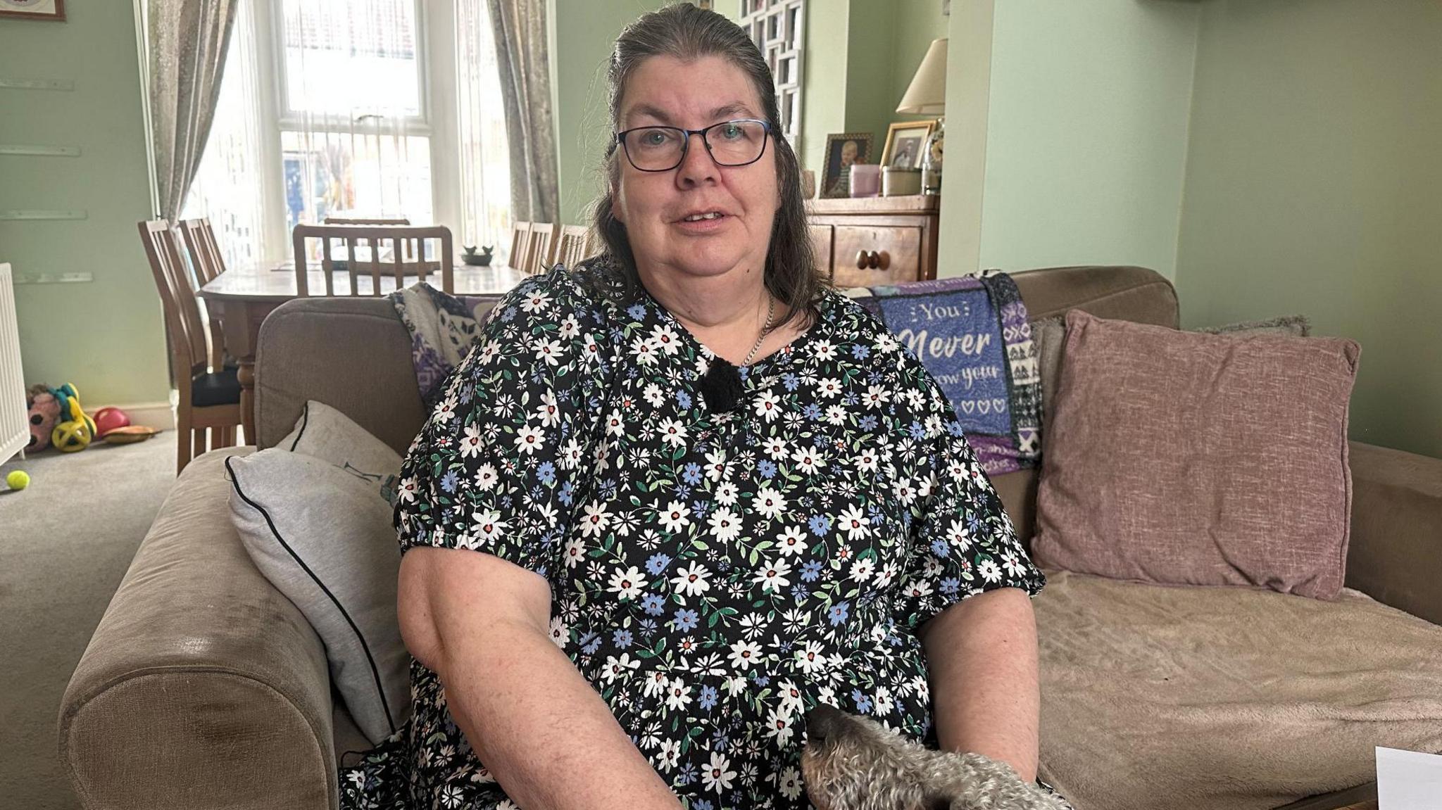 Nikki Paine pictured at her home in Ashford, Kent. She is sat on a beige sofa and has a dog at her feet. She is wearing a floral dress and has long brown hair and glasses.