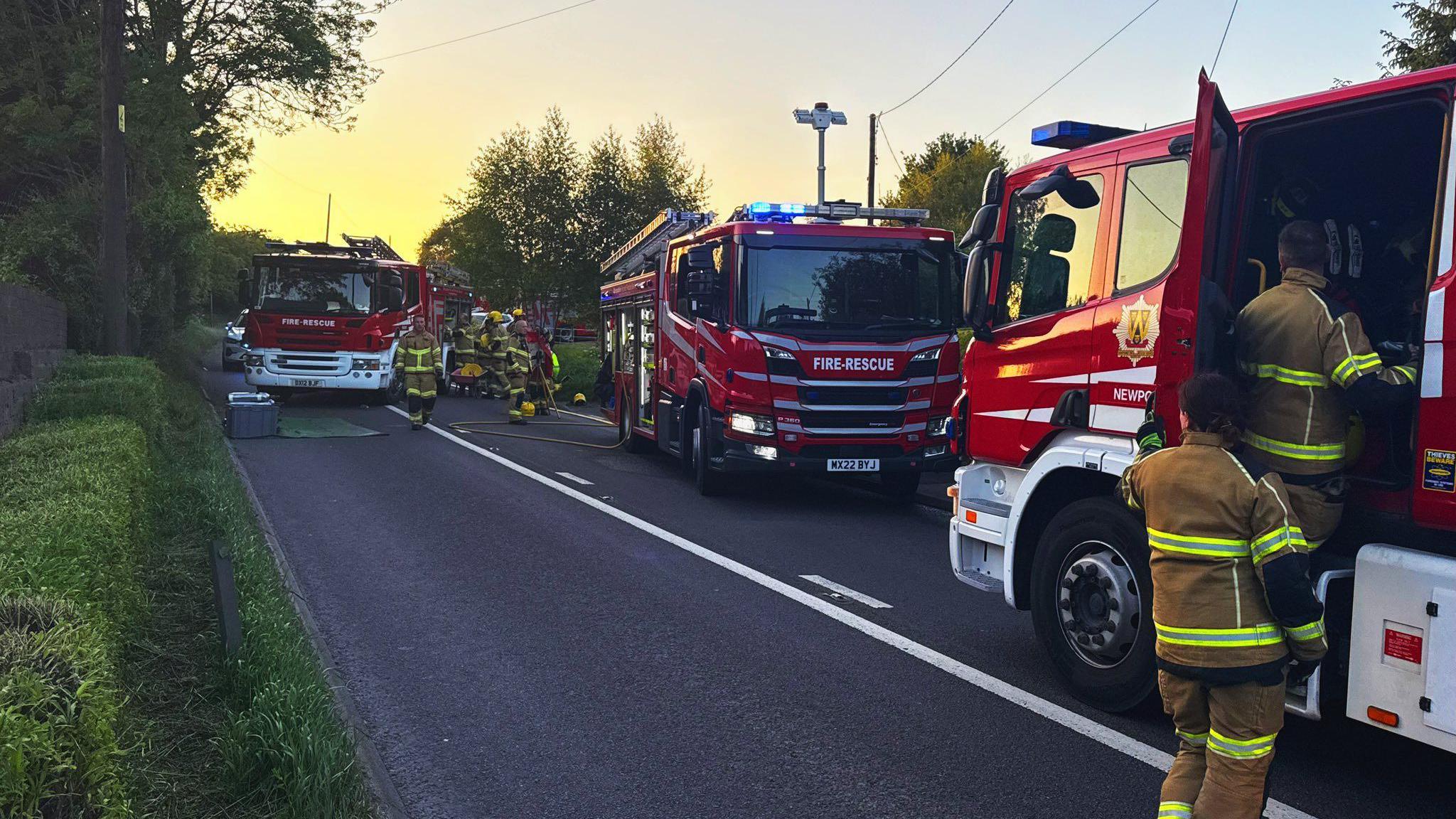 Fire engines are parked along a nearby road, blocking the carriageway. Firefighters wearing uniform are working around them.