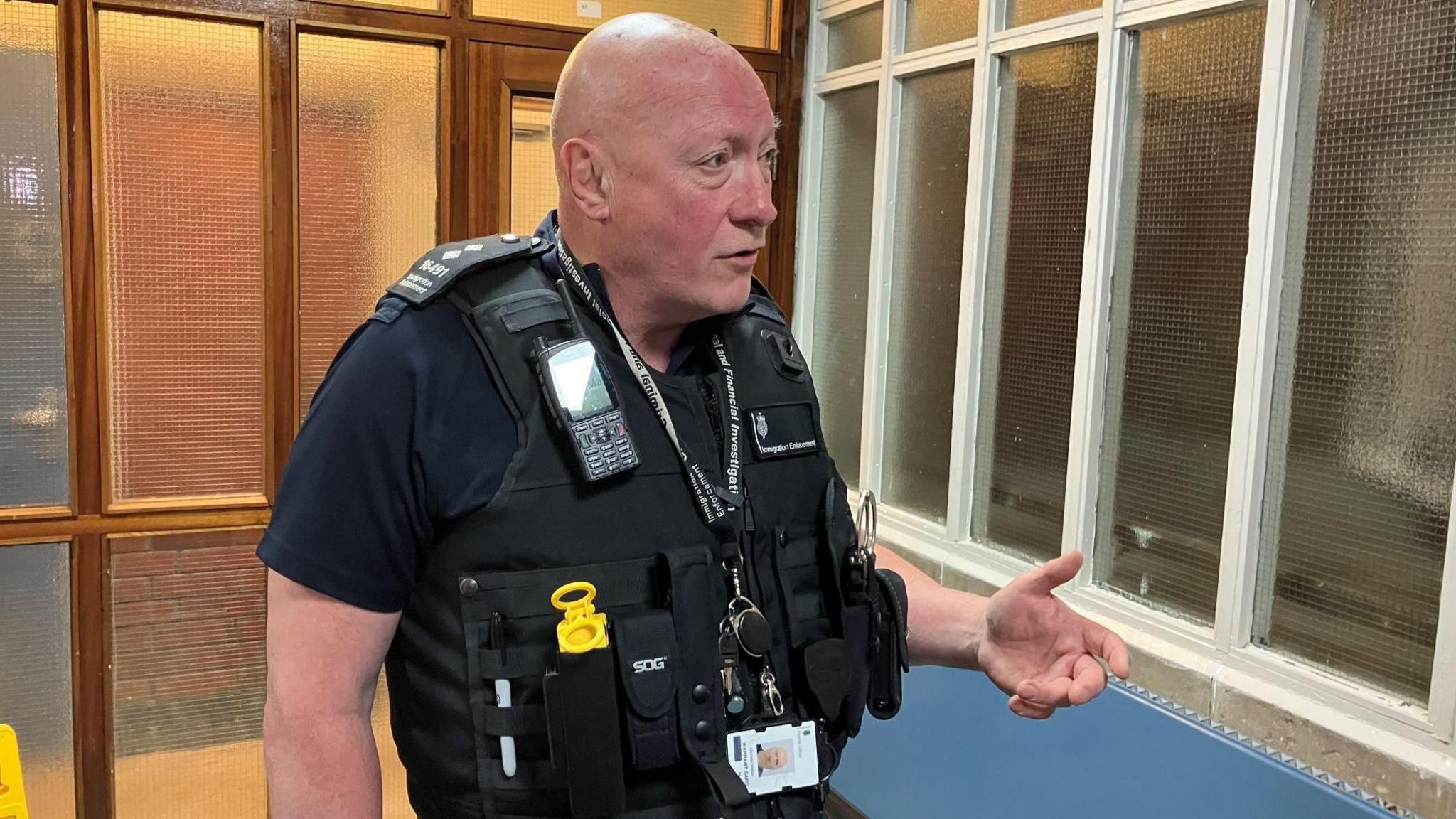 Chief immigration officer Mick Wilson gestures with one hand whilst he talks. He is wearing immigration enforcement uniform, which consists of a bullet proof vest with several pockets and a radio.