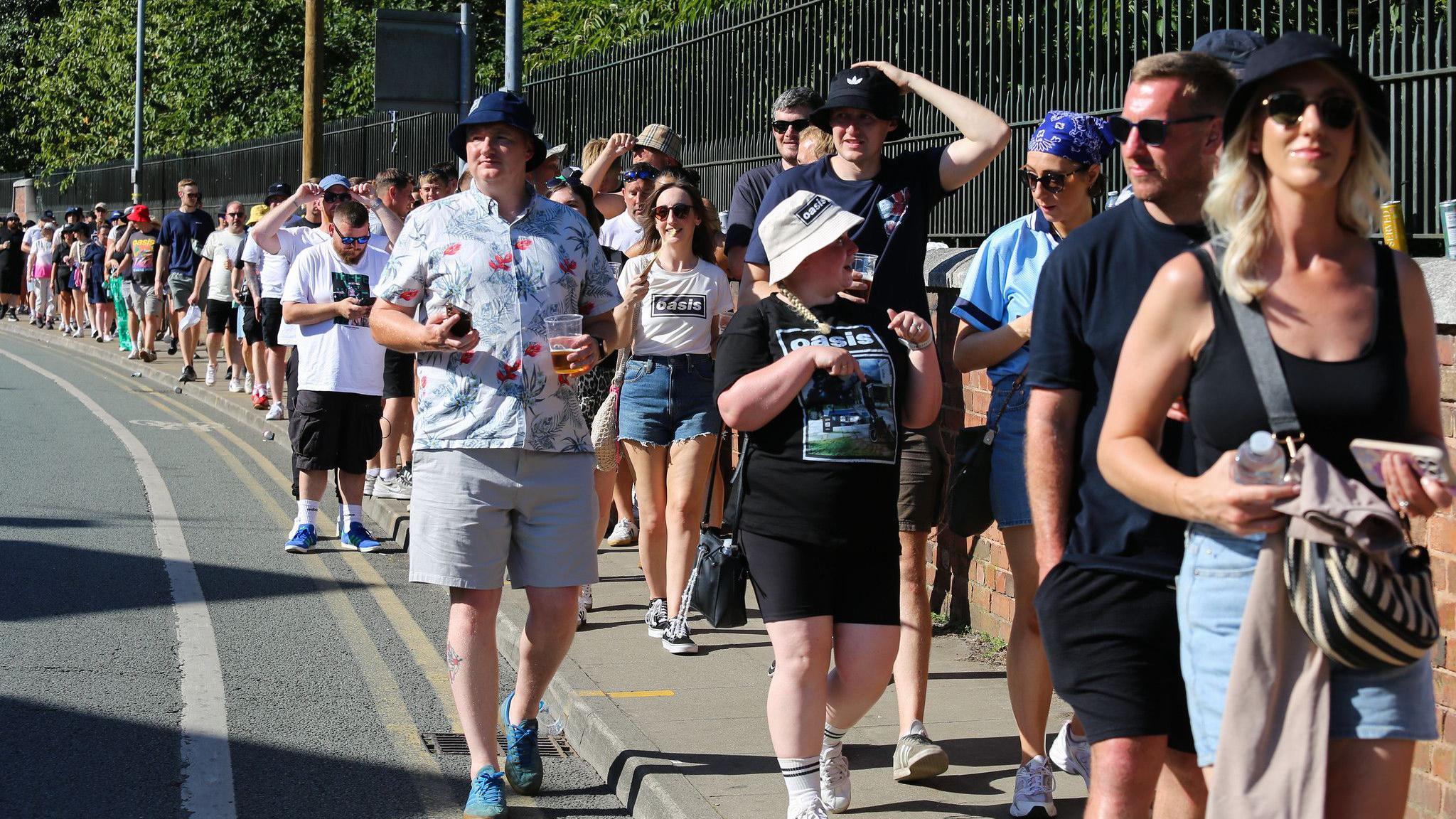 A long queue of Oasis fans in bucket hats, T-shirts and shorts walk beside the black iron railings of Heaton Park