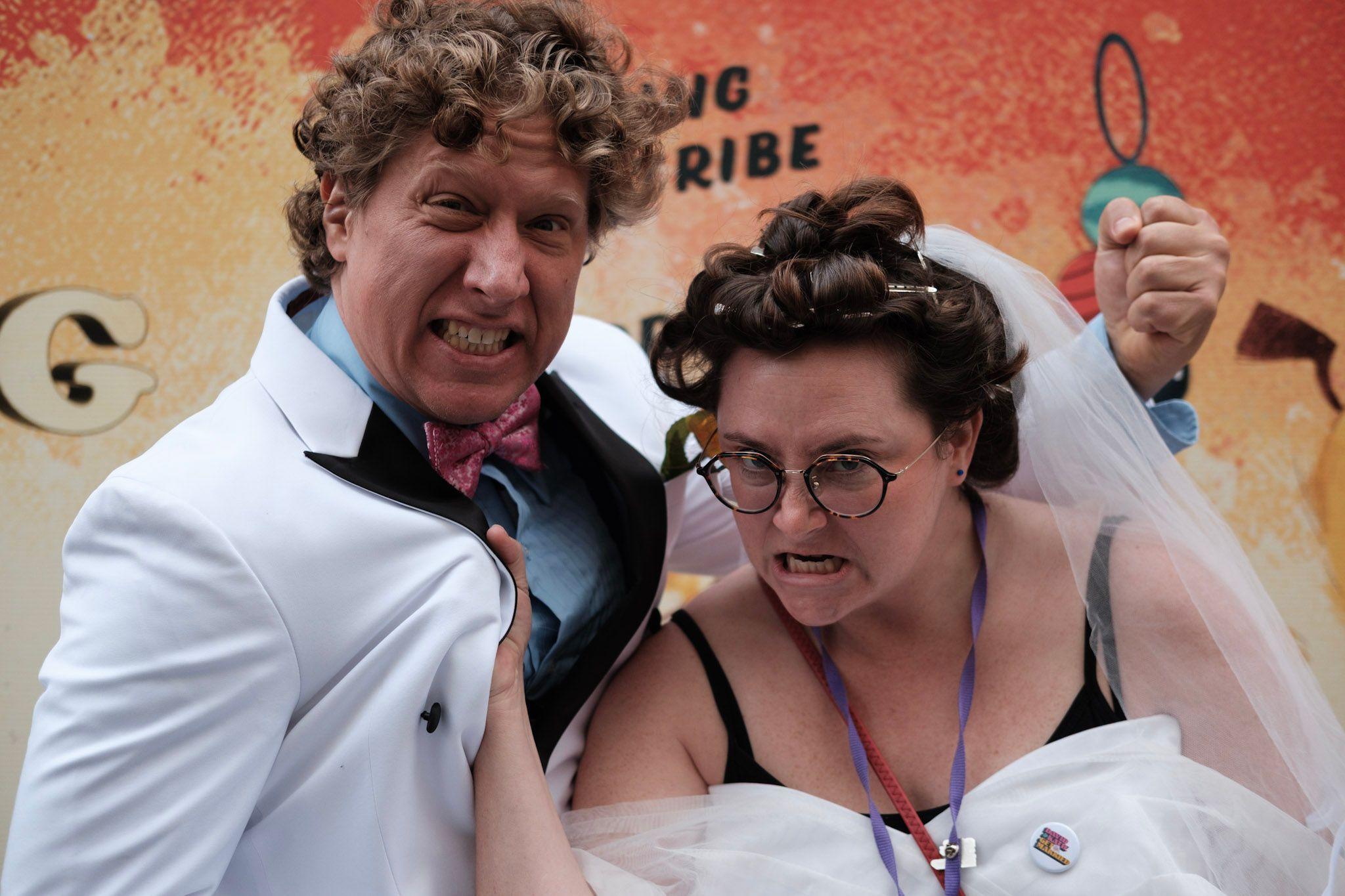 A man in a white suit and bow tie makes a fist beside a "bride" in a white dress, veil and with a grimace on her face as she grabs him by the lapels