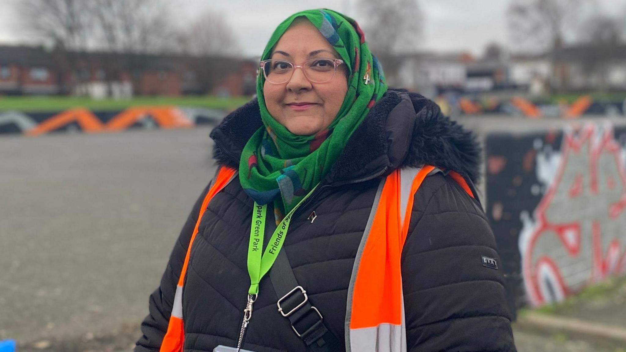 Sadia Khan is wearing a green head scarf and a black puffer coat. She is also wearing an orange hi-vis jacket and is standing in a park with a playground behind her