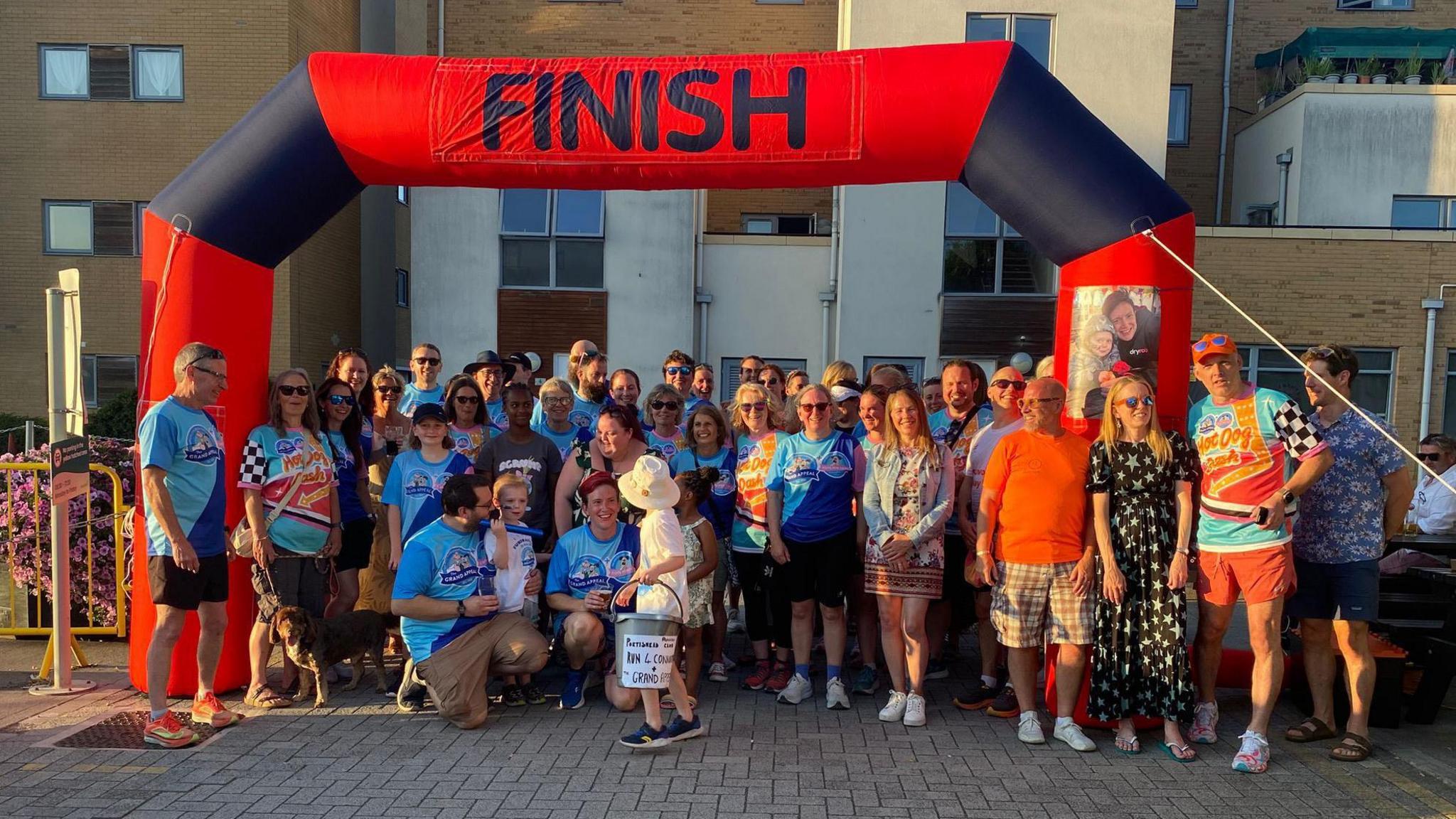 A large group of people standing below an inflatable red and black finish line. Many people are wearing blue Grand appeal branded t-shirts, and Connor can be seen in the foreground holding a grey donation bucket. Everyone is smiling and posing for a picture as the light turns orange at dusk.