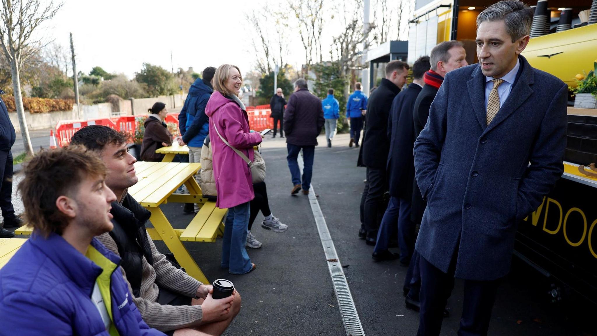 Fine Gael leader Simon Harris has greying hair and is wearing a navy suit coat, light shirt and tie and is standing against a food van. He is talking to two young men sitting at a yellow bench table