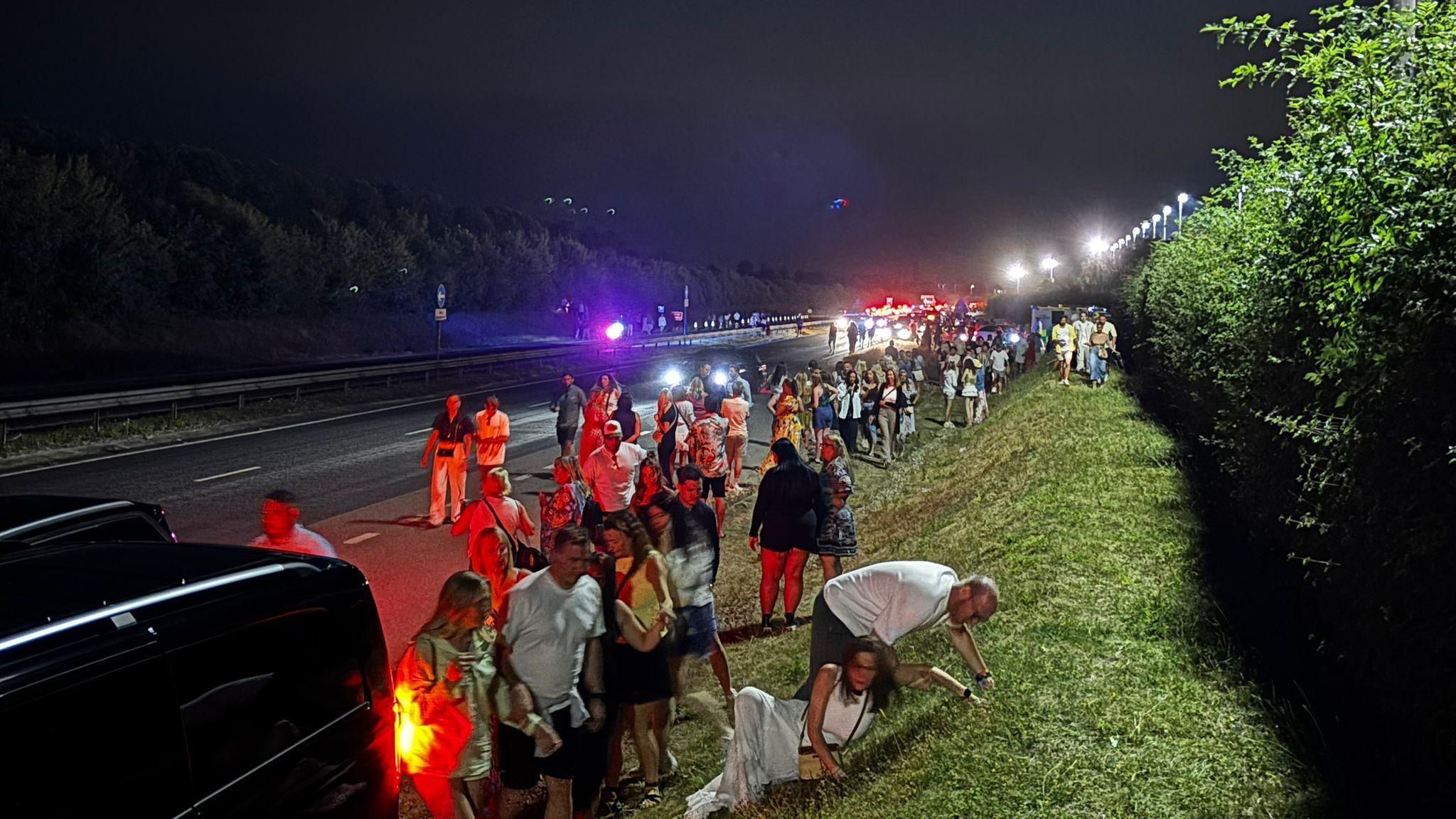 Dozens of people can be seen by a main road.  Standing on the verge and walking in the road. It is night time. Lights can be seen in the background.