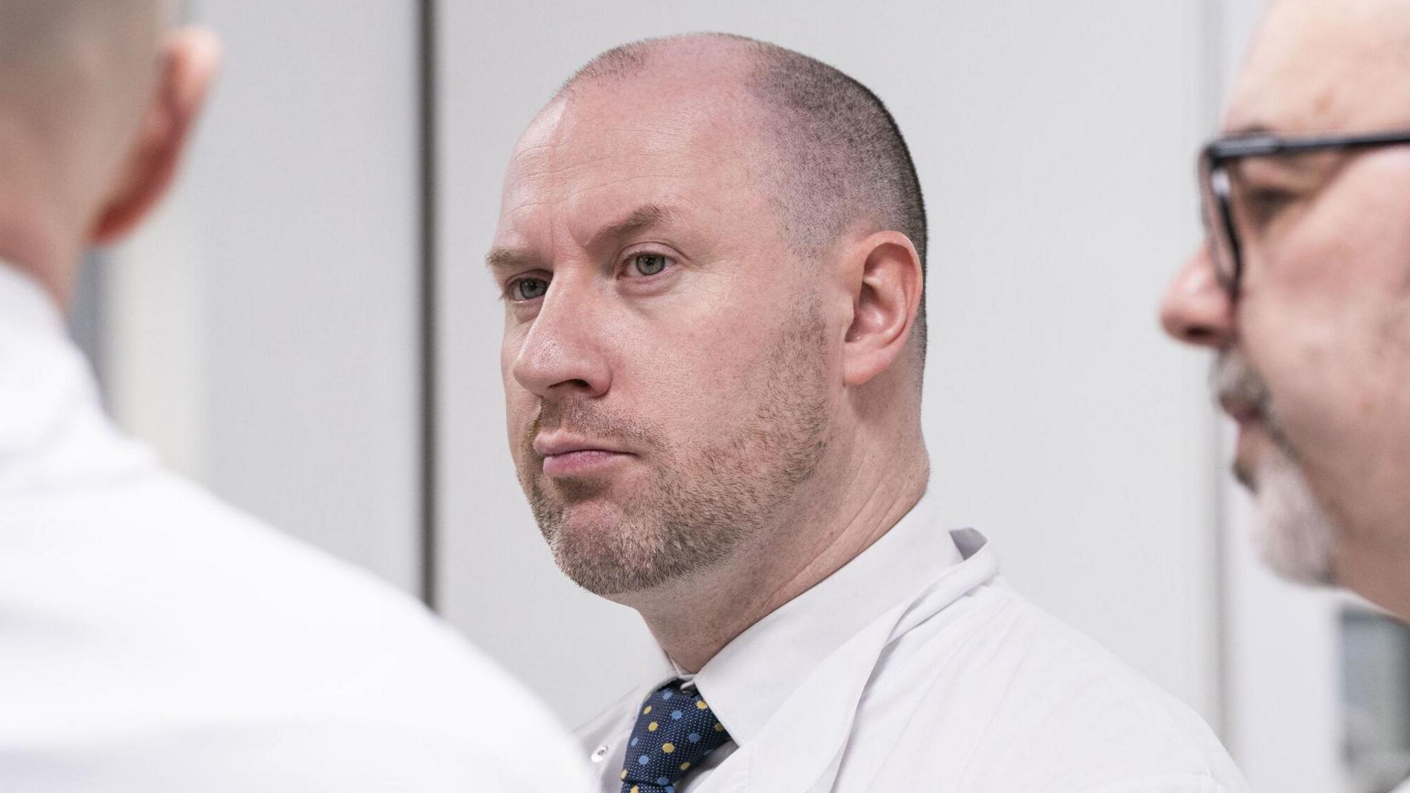 Neil Gray wears a white coat and shirt with a dark tie. He looks at a man in a white coat beside him