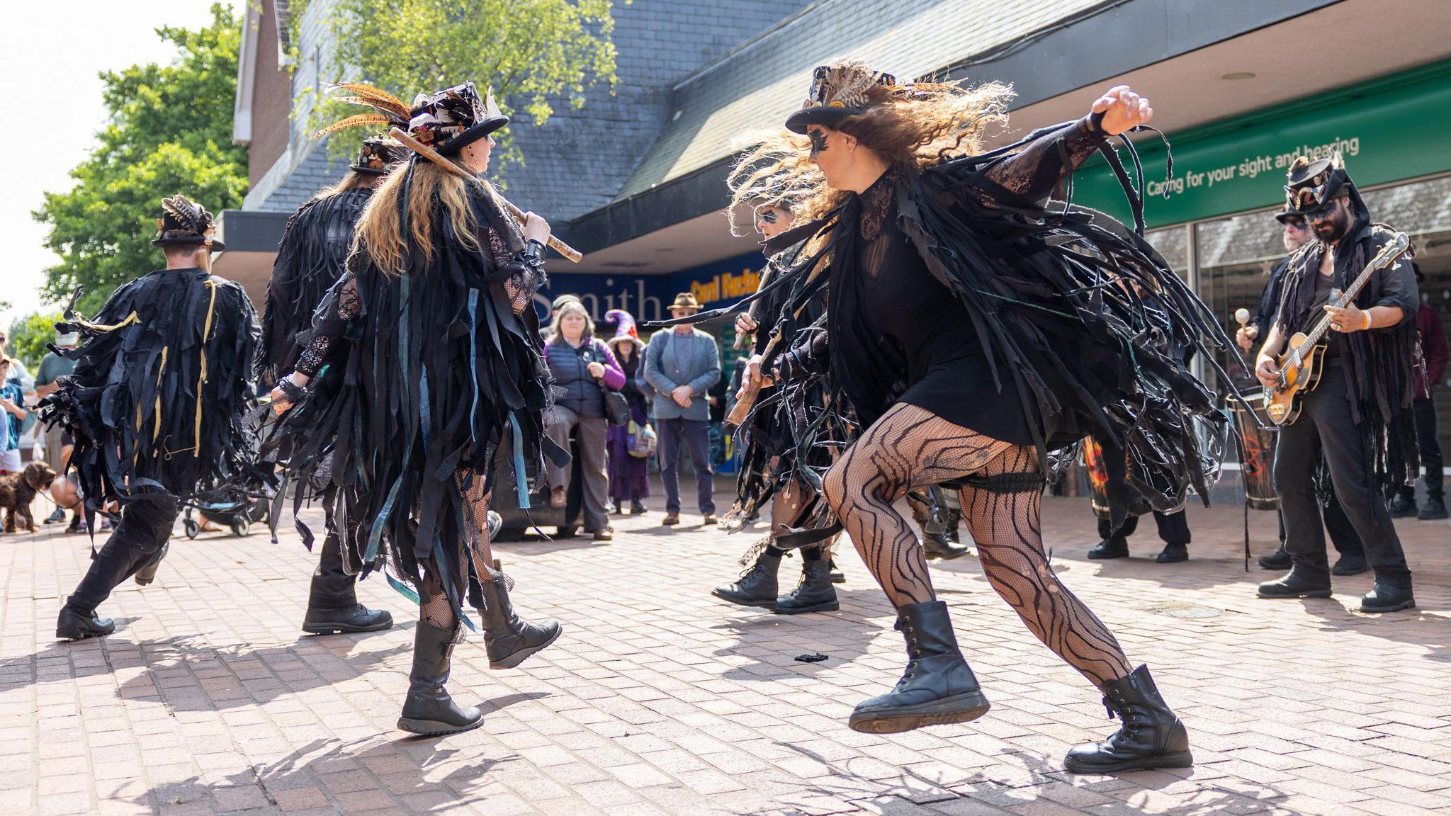 A group of morris dancers wearing all black with top hats and dancing around 