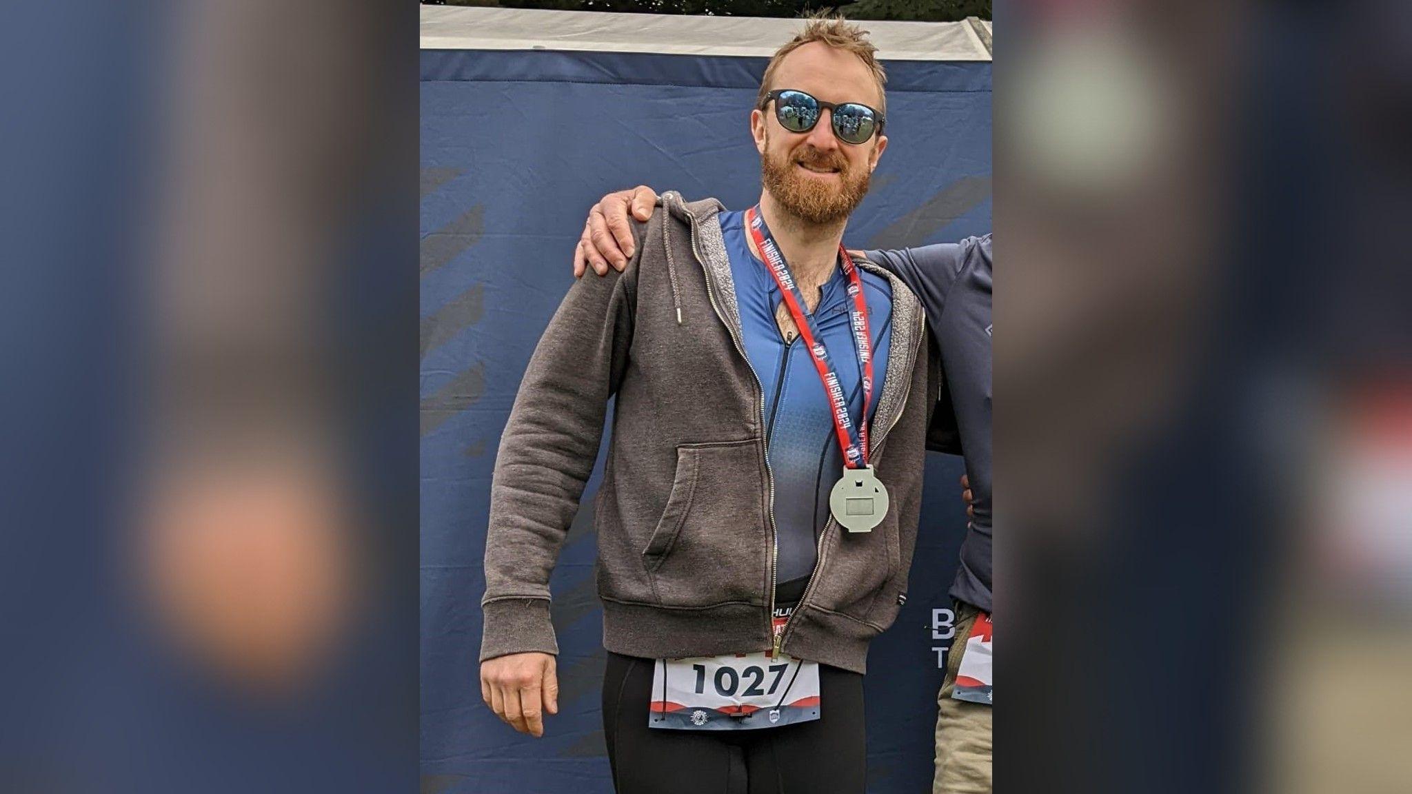 Dan with his medal after the triathlon, stood smiling at the camera with sunglasses on