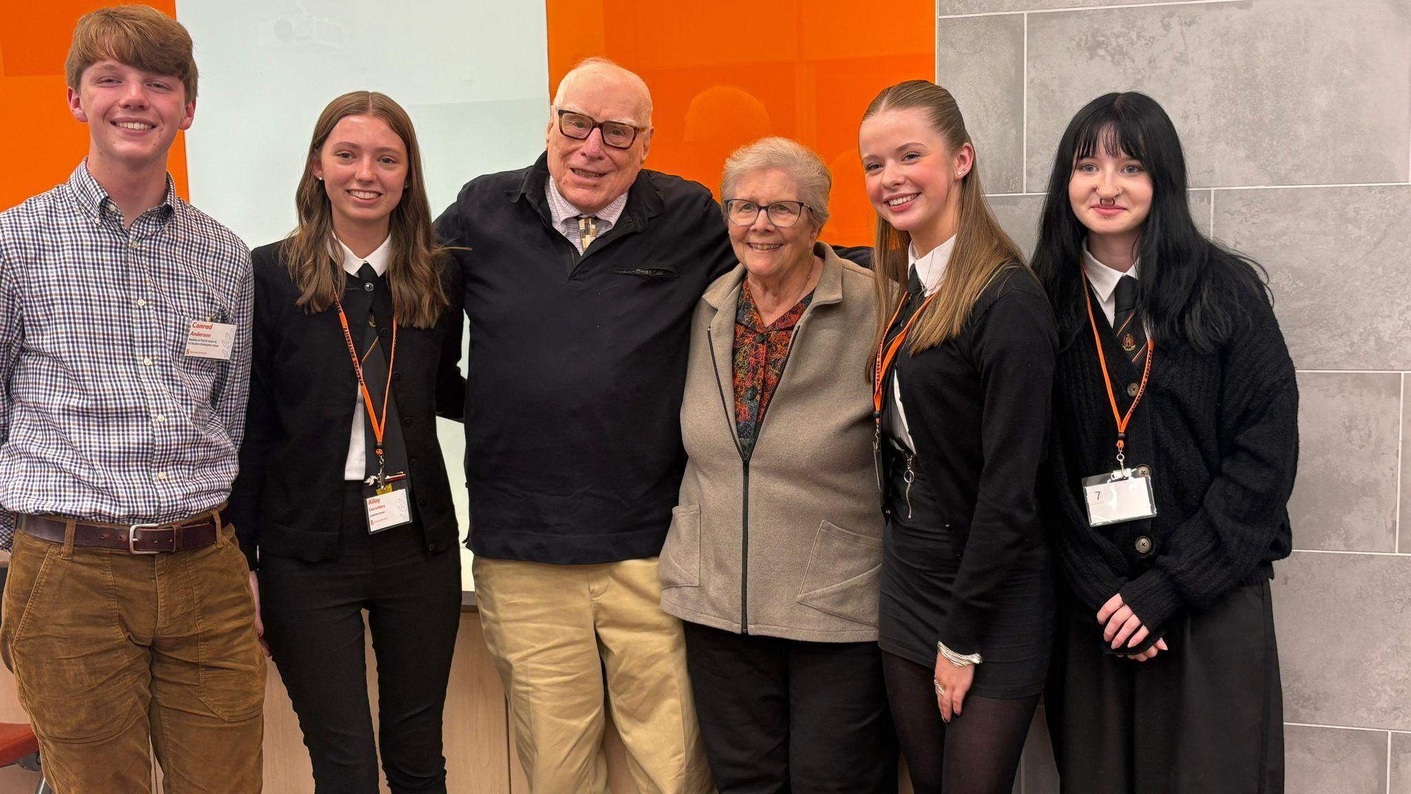 An older couple pose for a photograph with three young women and young man. The young women are all in school uniform.
