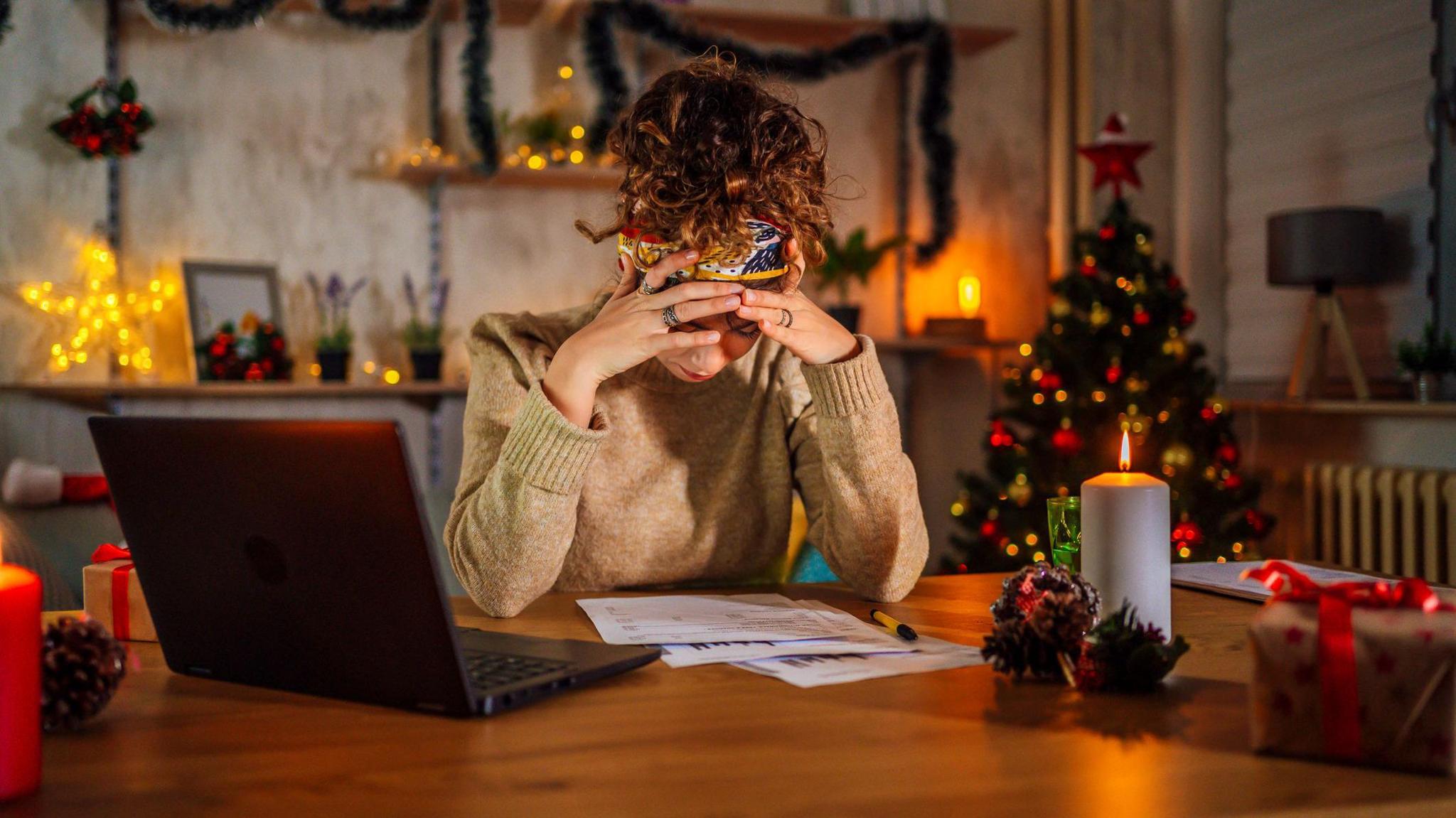 A woman has her hands resting on her forehead as she looks at bills on an oak table and a black laptop next to her. She has curly brown hair tied up on top of her head and is wearing a beige jumper. In the background, there is a Christmas tree and tinsel adorning shelves.