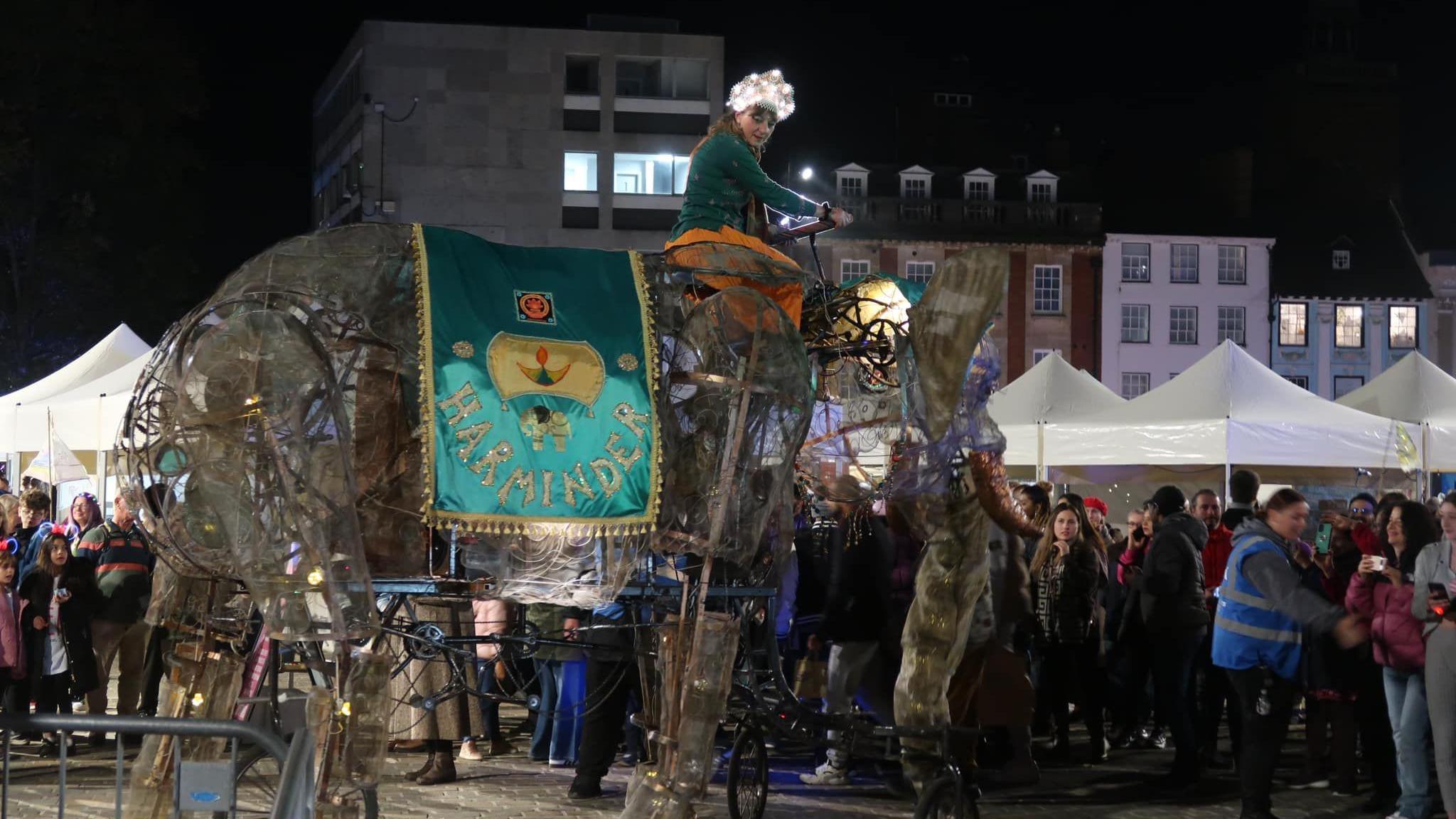 A mechanical elephant with a woman steering it on Northampton's Market Square, while revellers watch. Taken during the Diwali celebrations on 26 October 2024.