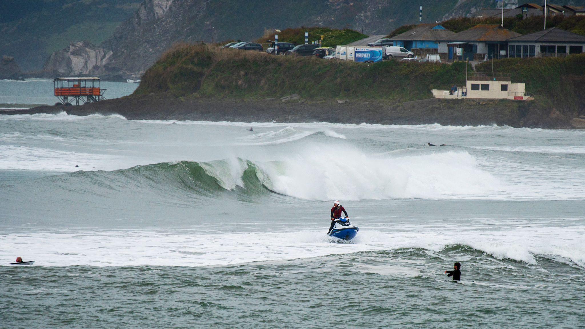 Noah Hindley on the blue and white Rescue Water Craft. He is located in the distance to the right at sea. In front is a swimmer, and a large wave breaking behind him. There is are grassy banks in the background with buildings located at the top, along with cars on the left. 