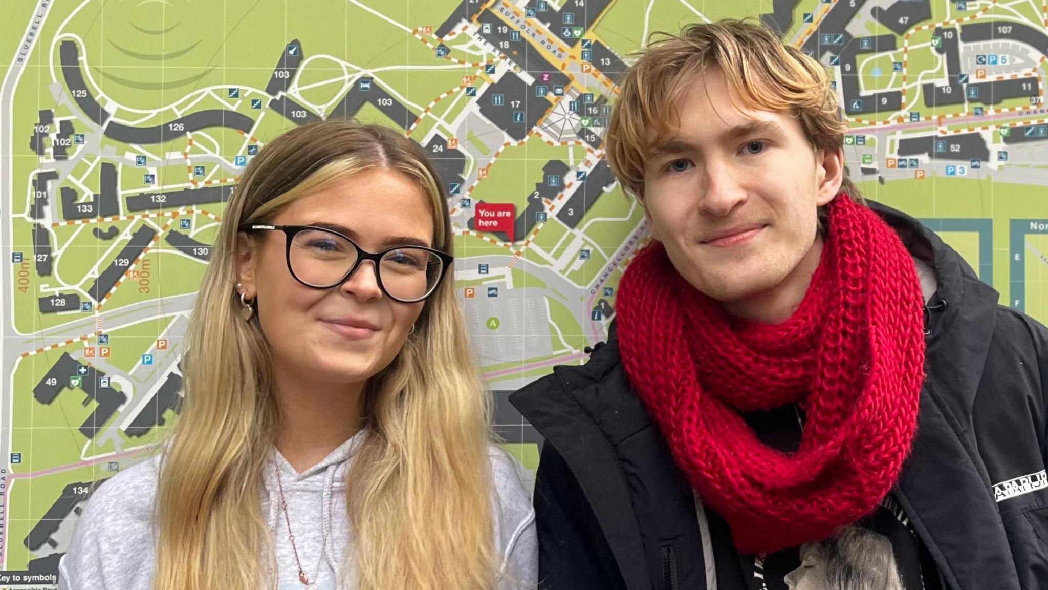 Grace Mahon and Charles Murphy, are standing side by side in front of a large, detailed campus map. Grace is wearing glasses and a light-coloured hoodie, while Charles is dressed in a black jacket with a bright red scarf. The map behind them shows buildings and pathways, indicating their location within the university grounds. Grace has long blonde hair and Charles has shorter blonde hair.