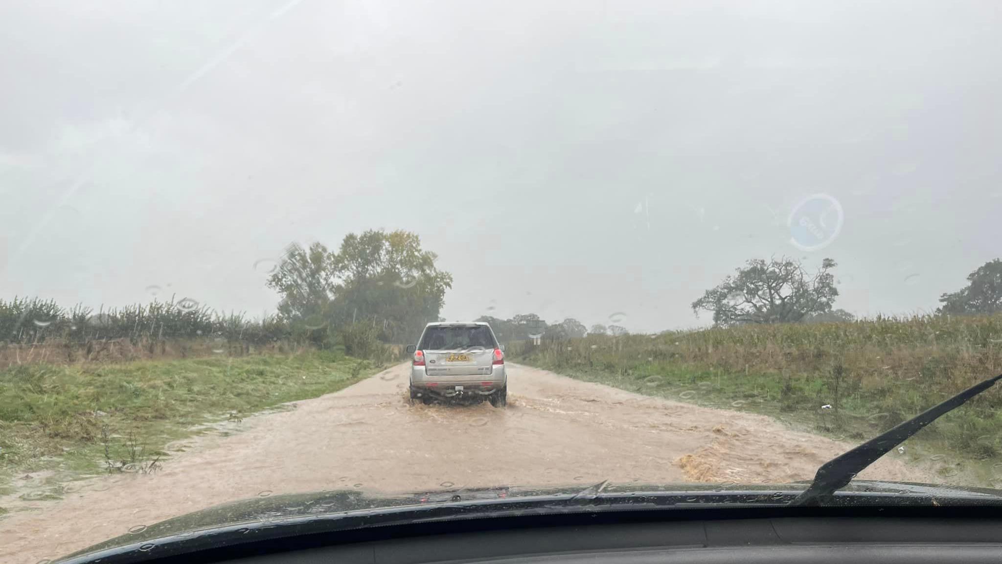 A wide view of a road from inside a car. There is a car driving through brown muddy floodwater which covers a road.