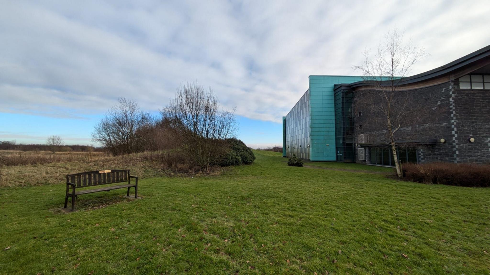 An area of grass to the side of a large grey and blue tile two-storey building. A wooden bench with a brass plaque is on the left and small shrubs/bushes are visible in the background.