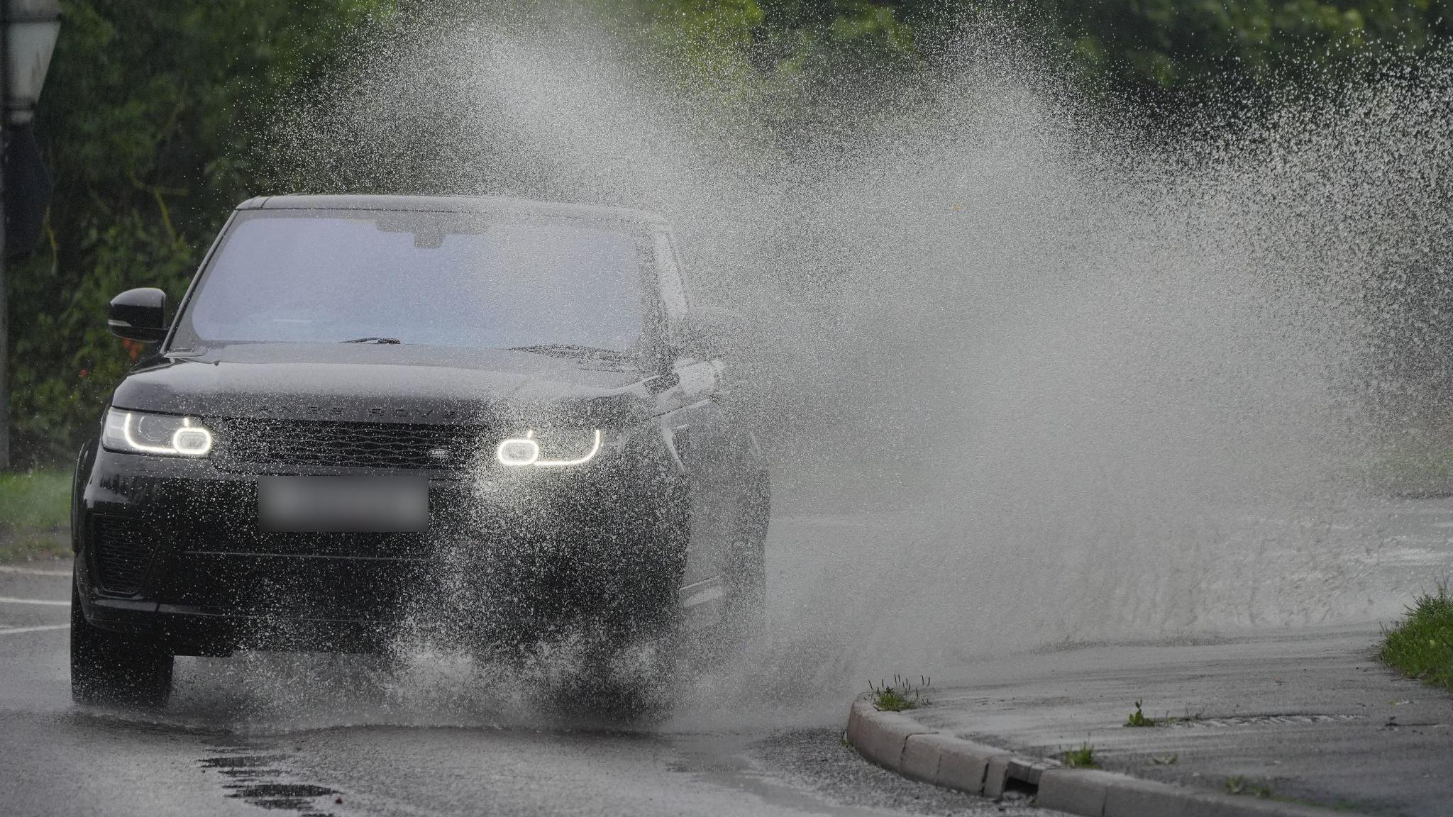 A black Range Rover drives through a large puddle on the side of a road during a rainy day, sending water flying high into the sky.