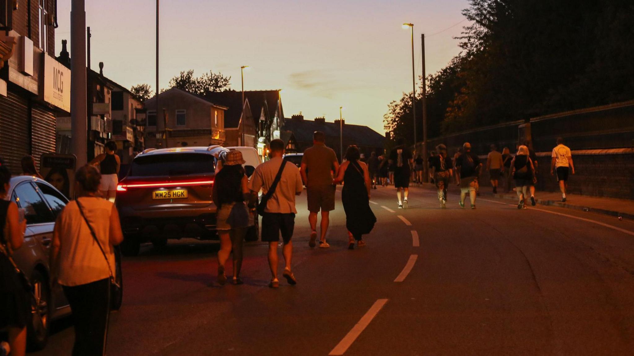 People in shorts and T-shirts walk up the road in the late evening light after Oasis at Heaton Park
