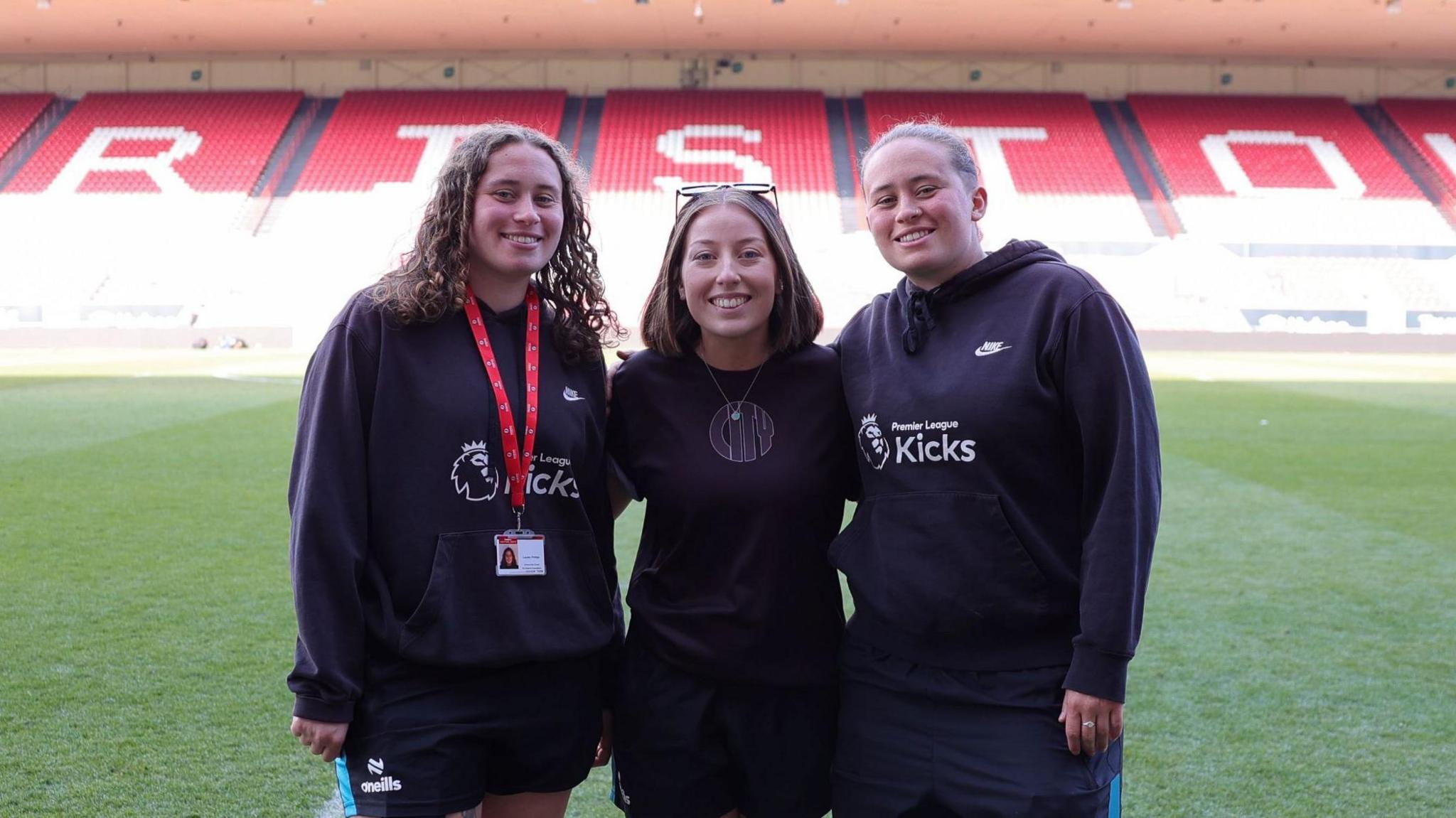 Three young women in black tops with white emblems stand on the pitch at Ashton Gate Stadium. In the background a stand with red and white seats is visible.