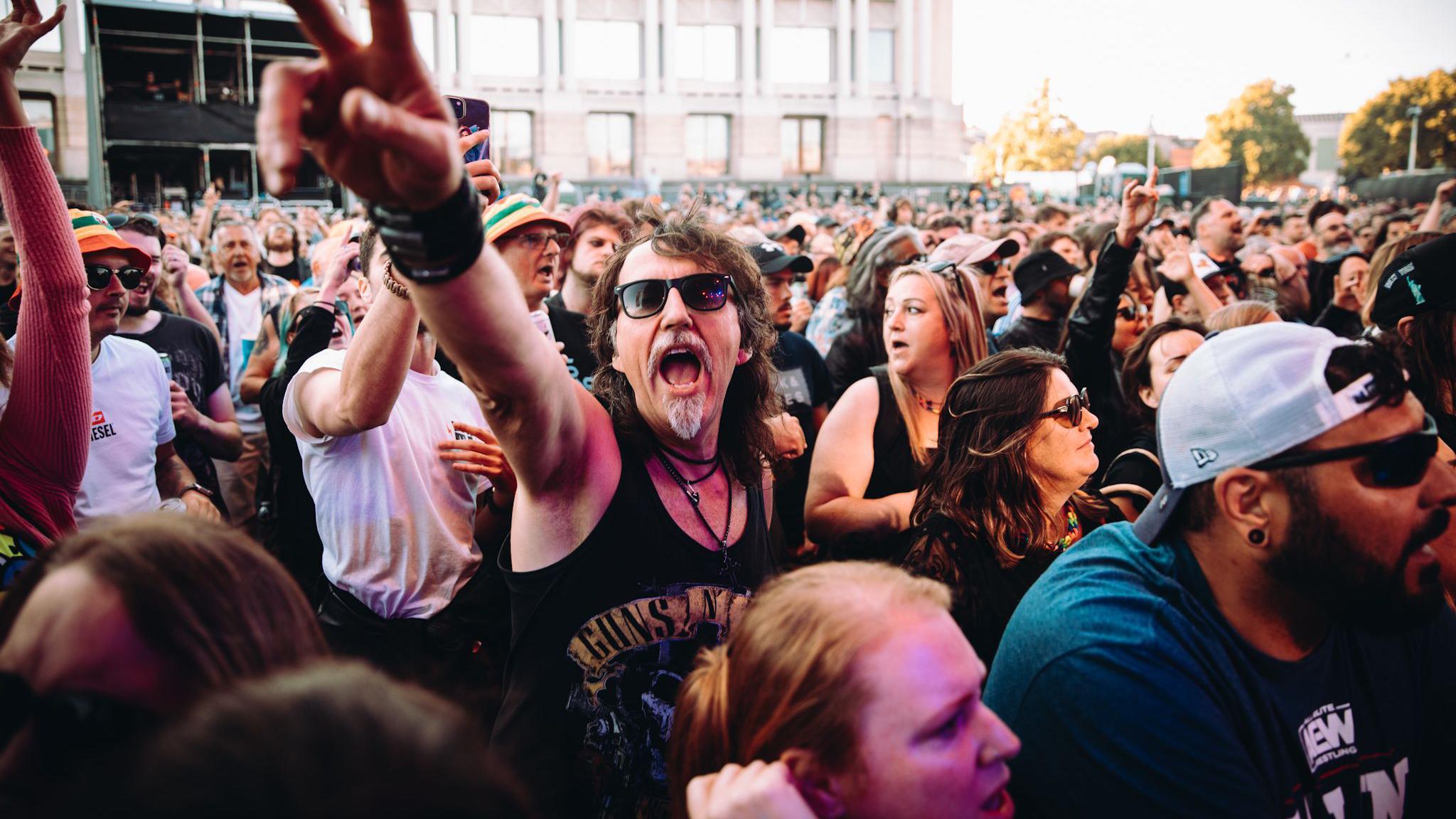 A large group of fans are packed into the Lloyds Amphitheatre during Bristol Sounds to watch the band Skindred. Many of them are looking towards the stage but one man is facing the camera and holding out an arm as he sings along. He has a black vest on, a goatee and moustache and sunglasses.