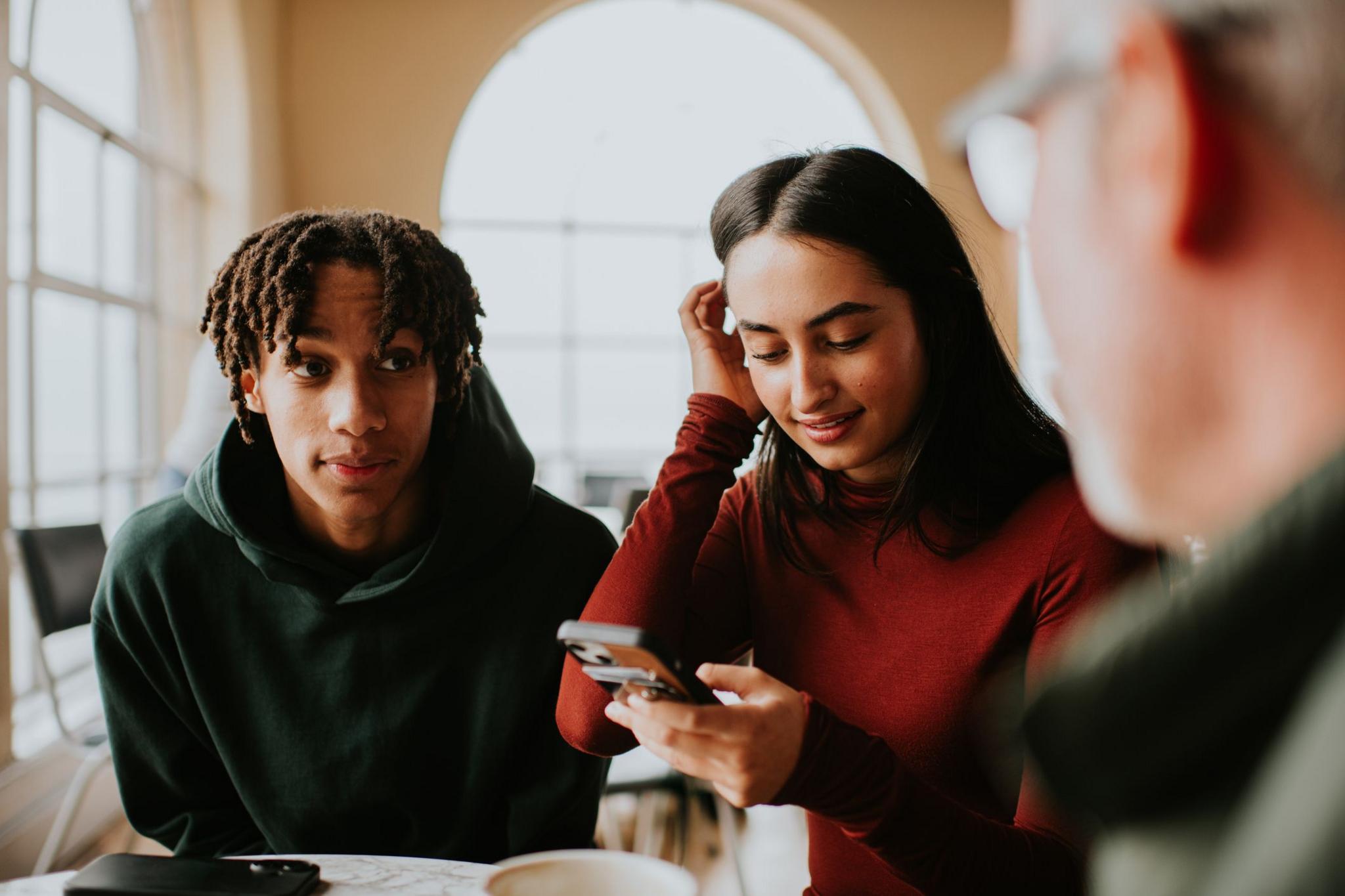 Two young adults sit at a table while one checks a smartphone, with an older man nearby