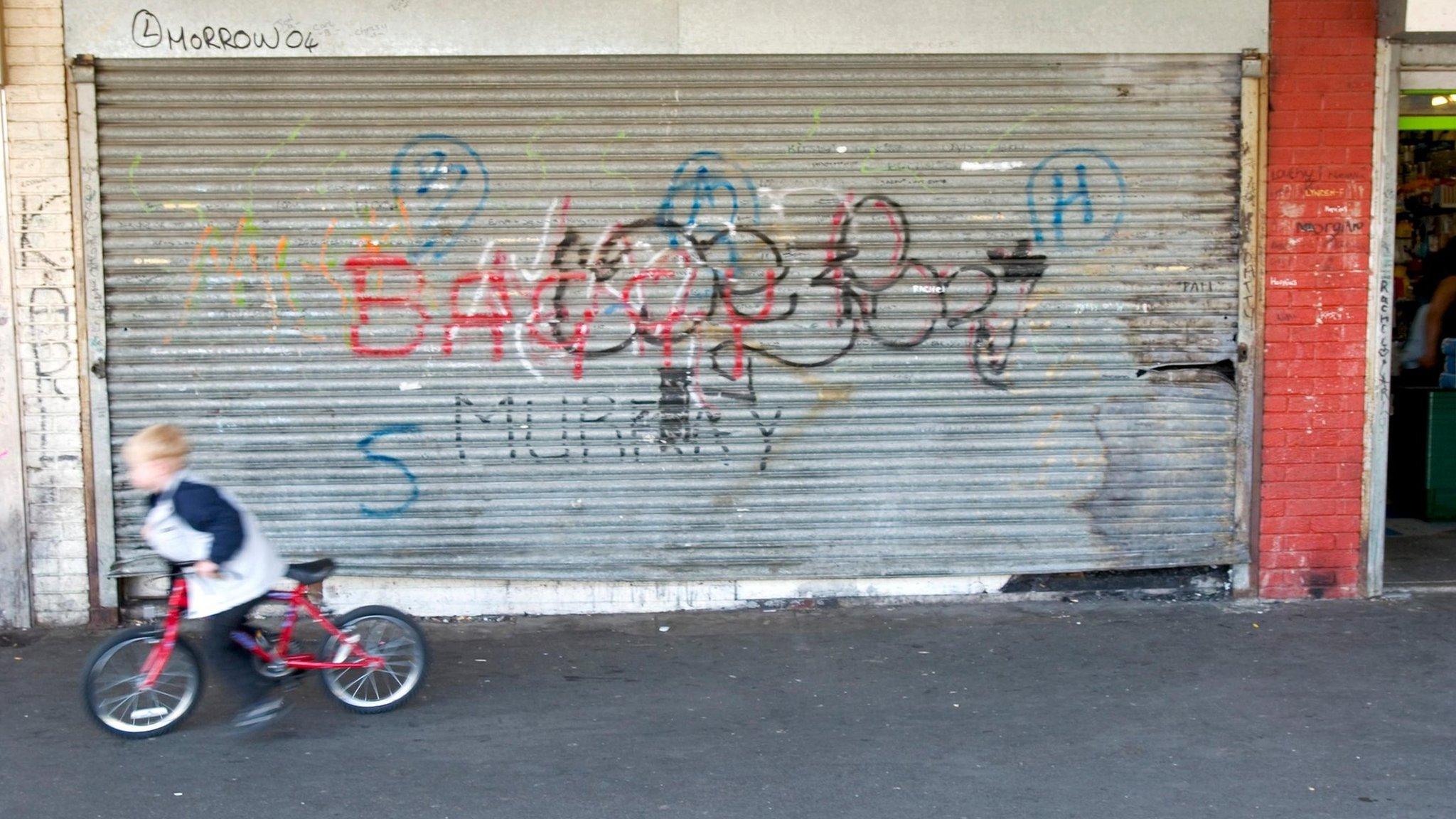 Boy on bike rides past boarded-up shop