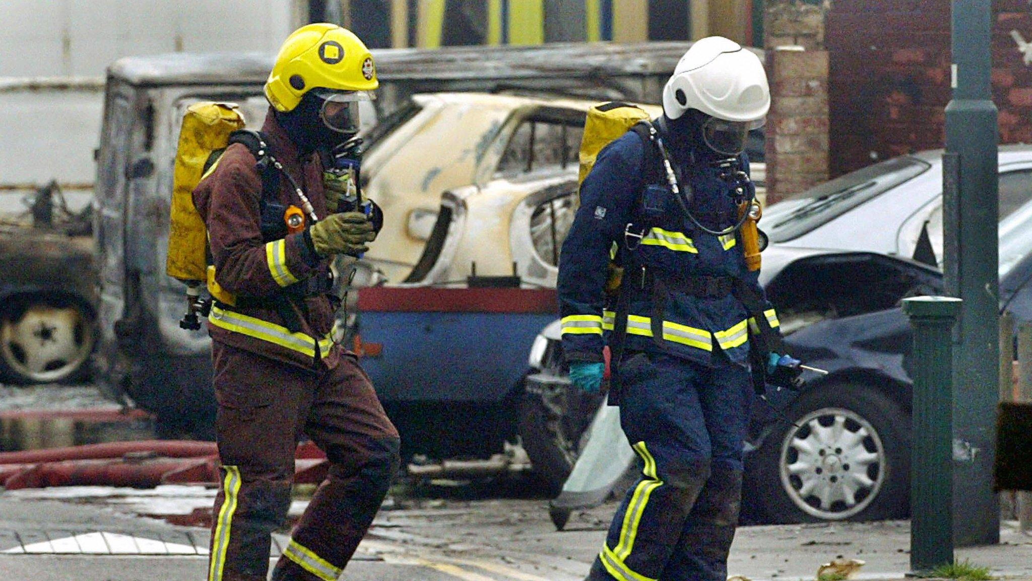 Firefighters walk past the ruins of a burnt-out car