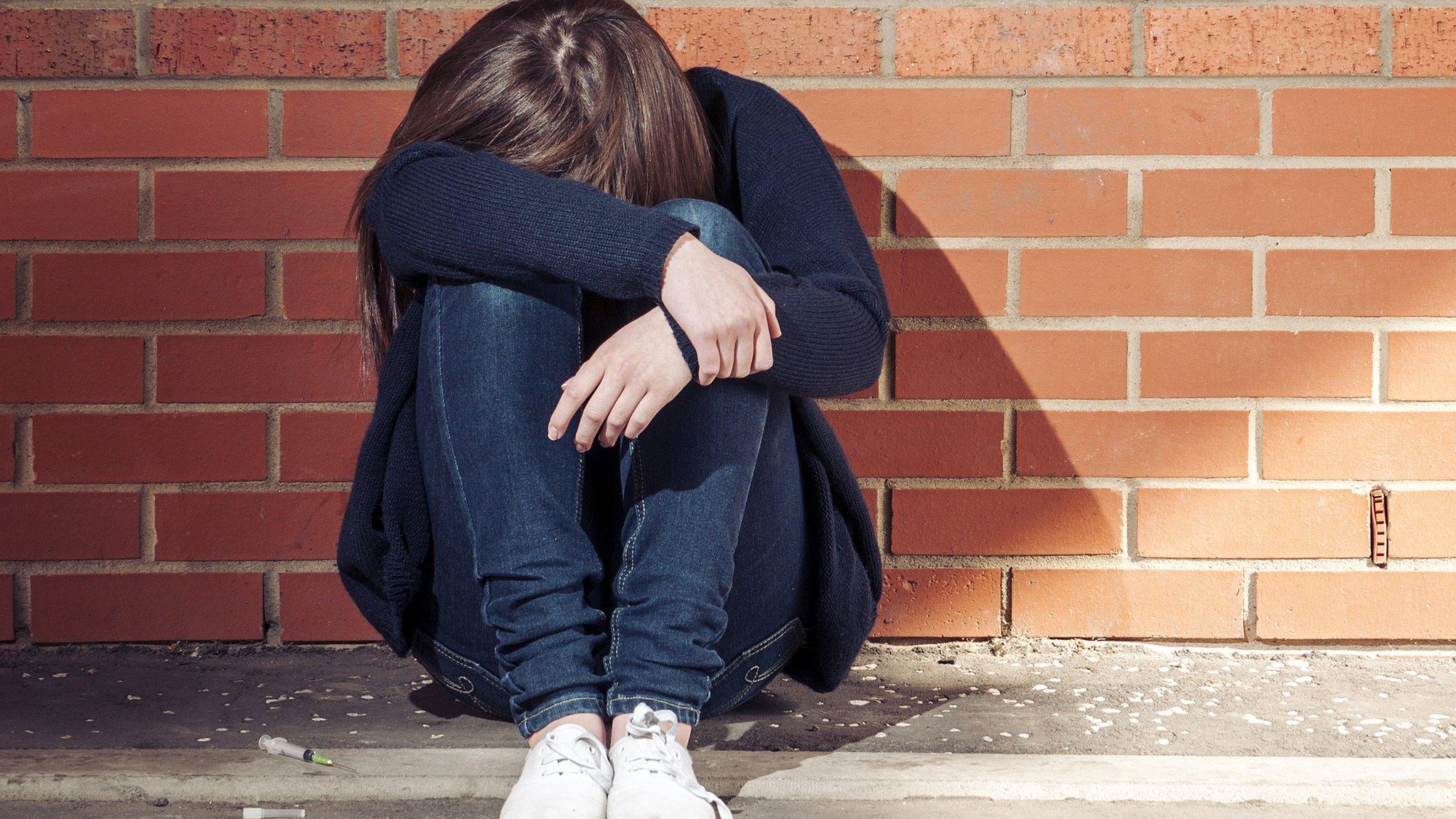 A woman sat against a wall with her arms over her face and her head in her lap