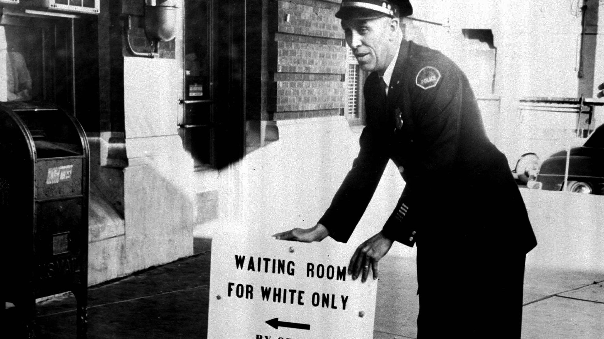 Policeman with a sign reading: "Waiting room for white only by order police dept."