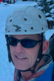 Close-up of a man wearing a white climbing helmet and dark sunglasses, standing outdoors in a snowy setting.