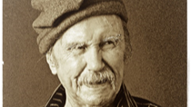 A black and white phot of Padraic Fiacc. He has a moustache and is wearing a woollen cap and a striped shirt.
