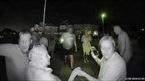 A still from a doorbell camera which shows a group of people all facing towards the house in a street. At the front is a woman holding hands with an older-looking lady