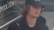 A CCTV image showing a young man with brown curly hair emerging from under a black baseball cap. He is wearing a black Monterrain hoodie