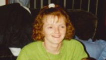 The late Tracy McCormack with bobbed curly brown hair in a ponytail wearing a lime green jumper holds twin babies in her arms. She is sitting on a sofa with blue cushions. She is smiling.
