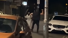 Two men in a road fighting with large blades. One is wearing all black and the other is wearing all grey with a balaclava. There are cars parked on the side of the road and it is night time.