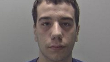 Mugshot of Alexander Jesus, wearing a blue t shirt. He is against a grey backdrop and has short brown hair. He is looking straight into the camera
