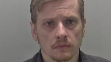 Mugshot of a man with short blonde hair, in a side part with a short fringe. He has a moustache and beard and is wearing a grey shirt.