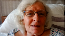 Annette Smith lying in bed slightly propped up on a white pillow, wearing oval glasses and a nightwear top with blue hearts on. She has wave white hair.