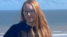 Cassidy standing outdoors on a sunny day with the sea and a sandy shoreline visible in the background. She is wearing glasses, a black zip‑up jacket and matching black trousers. Her long, ginger hair falls over one shoulder.