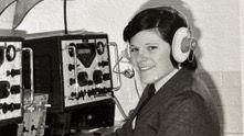 A grainy black-and-white archive photo shows a young Pádraigín smiling at the camera behind a large machine on a desk covered in dials and switches. She is wearing a suit and tie and has a large headset on her head.
