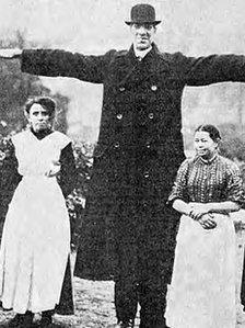 English Giant Fred Kempster standing next to two women of normal height